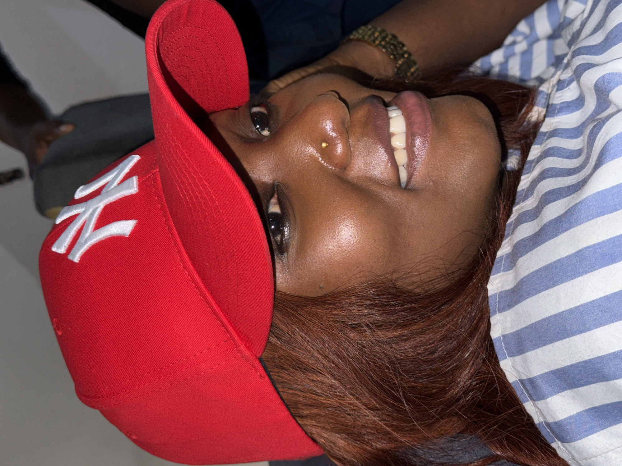 A close-up portrait shows a Black woman wearing a red baseball cap with a white logo, possibly the New York Yankees logo. She has brown hair and is wearing a blue and white striped shirt. A small gold stud is visible in her nostril. The location cannot be determined from the image. No specific actions or interactions are depicted.