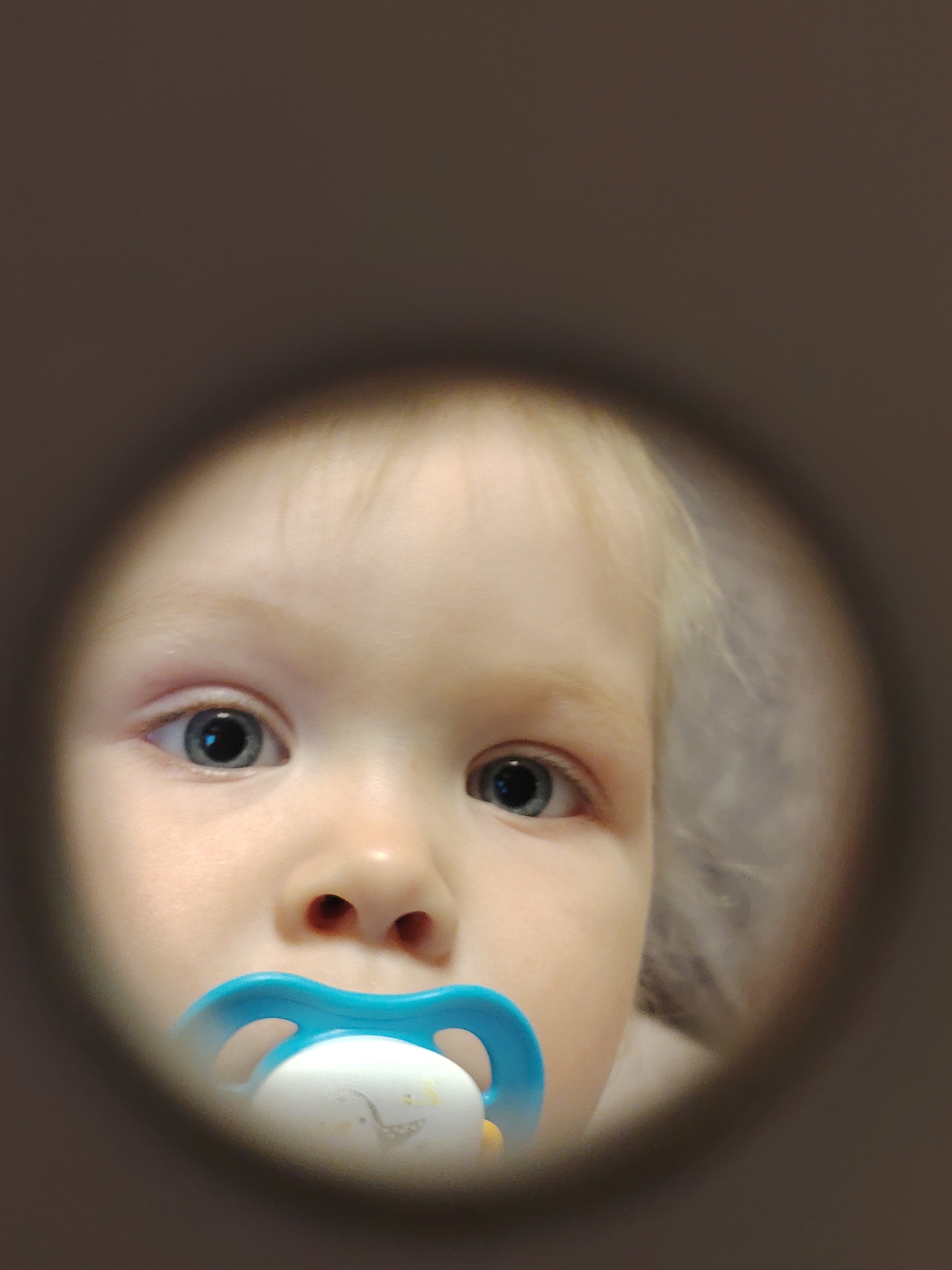 A close-up, eye-level shot shows the face of a young child with light skin, framed by what appears to be a circular hole or opening. The child has fair hair and blue eyes. A blue and white pacifier is in the child's mouth. The background and the immediate surrounding area of the circular opening are out of focus and appear to be a solid, muted color. The image lacks specific details to determine the location in Chaikovsky, Russia.
