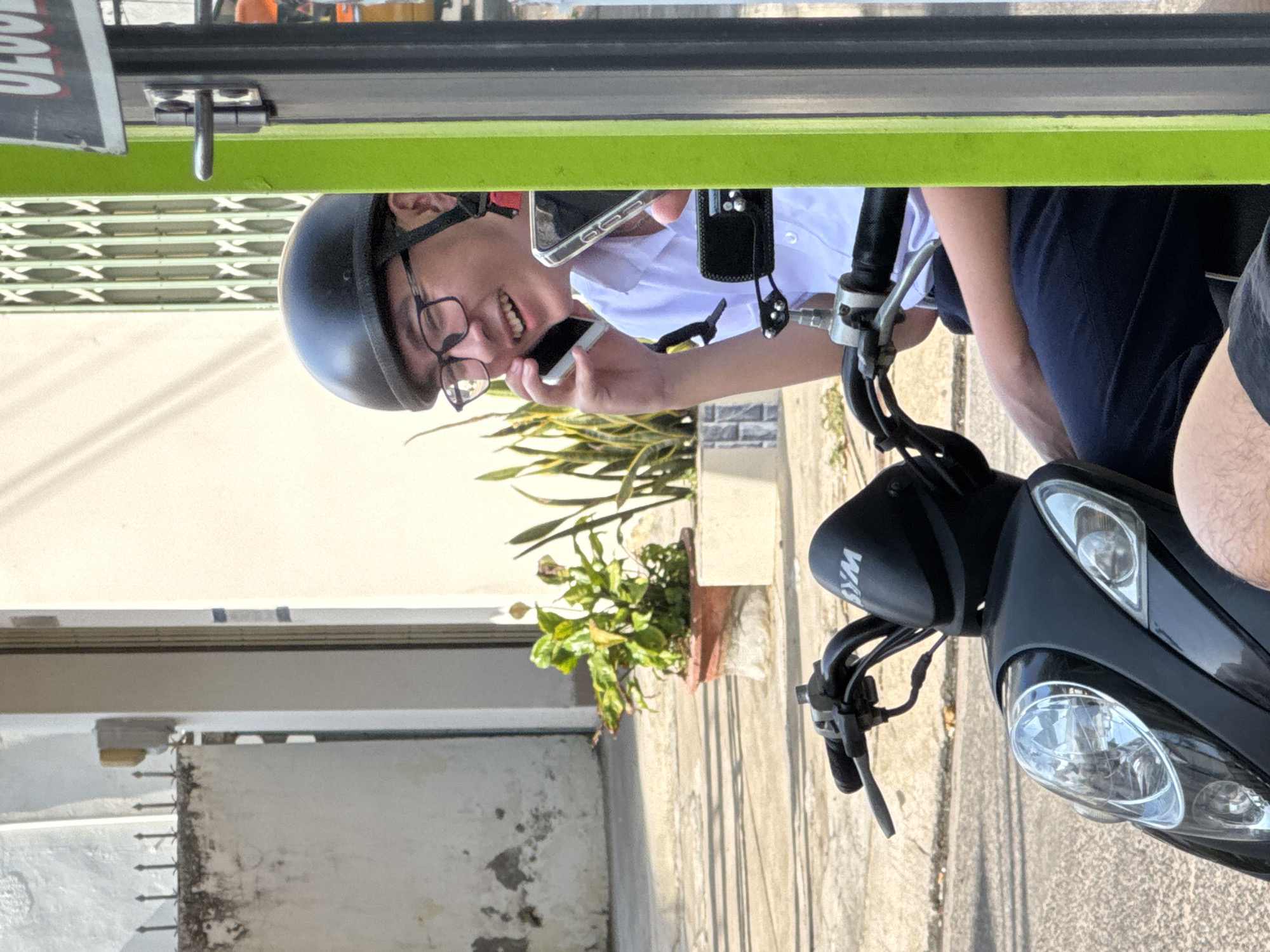 A young Vietnamese man wearing a black helmet and glasses is riding a black scooter. He is holding a white smartphone to his right ear and appears to be speaking on it. He is also holding another smartphone in his left hand, with its screen facing outwards. The man is wearing a white collared shirt and dark blue pants. He is on a street with a light-colored pavement and buildings in the background. To the right of the man is a bright green pole and a grey pole. The image appears to have been taken in the city of Nha Trang, Vietnam, based on the context provided.