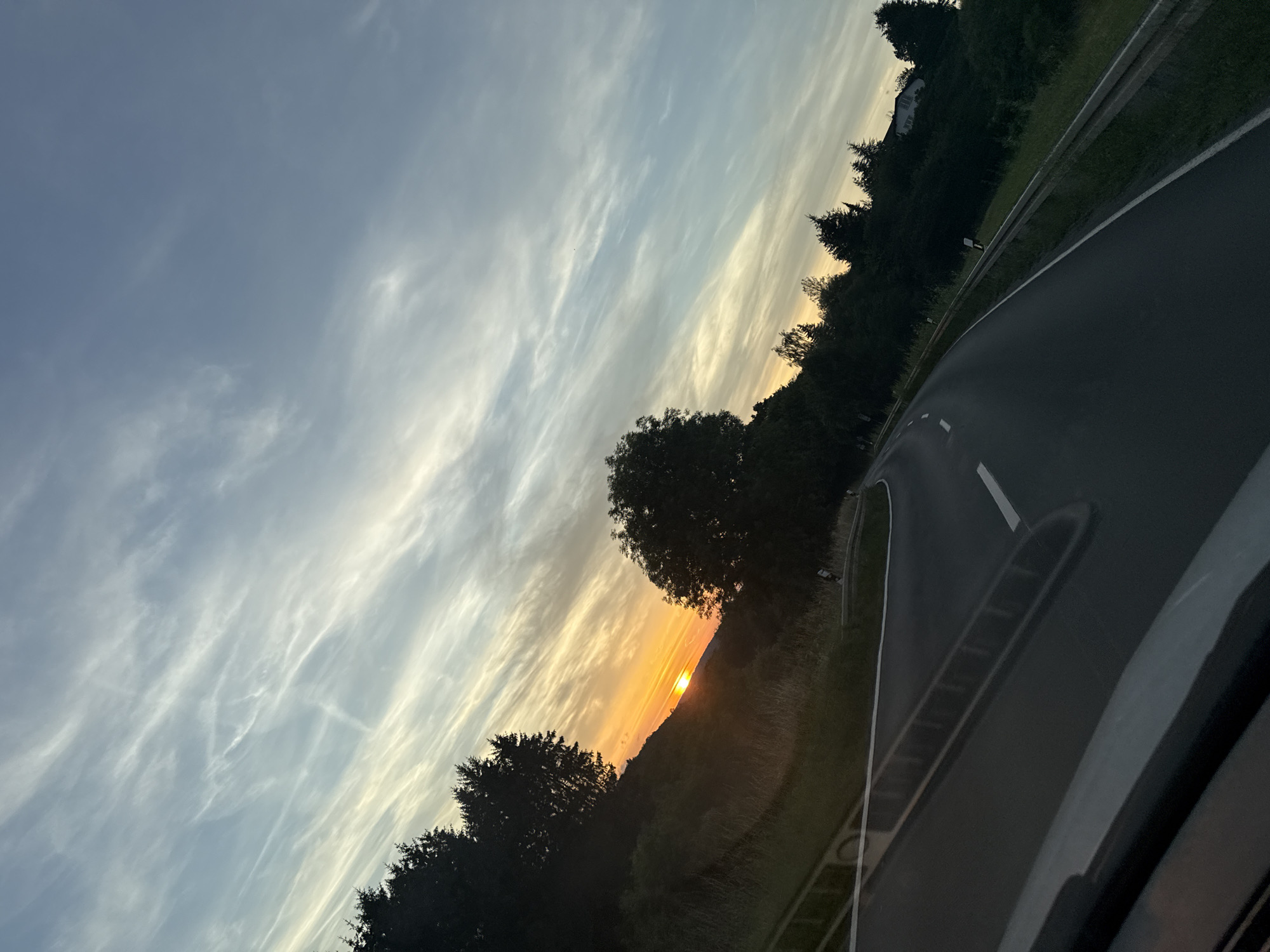 The image shows a sunset view from a vehicle traveling on a winding road near Kröffelbach, Germany. The sun is low on the horizon, casting a warm orange glow through streaks of white clouds in a pale blue sky. The road, with a white dashed line marking its center, curves through a landscape of rolling hills covered in dark green trees and grass. A guardrail runs alongside the road, and on the right side of the road, a house is visible nestled among the trees on a hillside. The foreground shows a portion of the vehicle's interior, including the dashboard and windshield.
