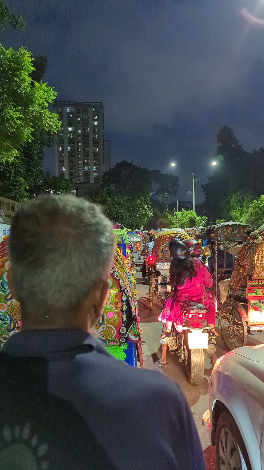 The image depicts a traffic jam in Dhaka, Bangladesh, at night. The scene is characterized by a dense collection of colorful rickshaws, illuminated by streetlights and the glow from a tall building in the background. In the foreground, the back of a man's head and shoulders are visible, suggesting he is a pedestrian observing the traffic.
The rickshaws are ornately decorated with vibrant patterns and designs, typical of transportation in Dhaka. One rickshaw in the middle of the frame carries two people: a woman in a pink outfit riding on the back of a motorcycle that is attached to the rickshaw, and another person, partially obscured, sitting in the rickshaw itself. The motorcycle's taillight is on, and a license plate is visible.
Other rickshaws are seen in front and behind, forming a long line. The street appears to be paved, and trees line the side of the road. The sky is dark and cloudy, with some ambient light filtering through, possibly from the city. The overall impression is a bustling urban environment during a nighttime traffic congestion.