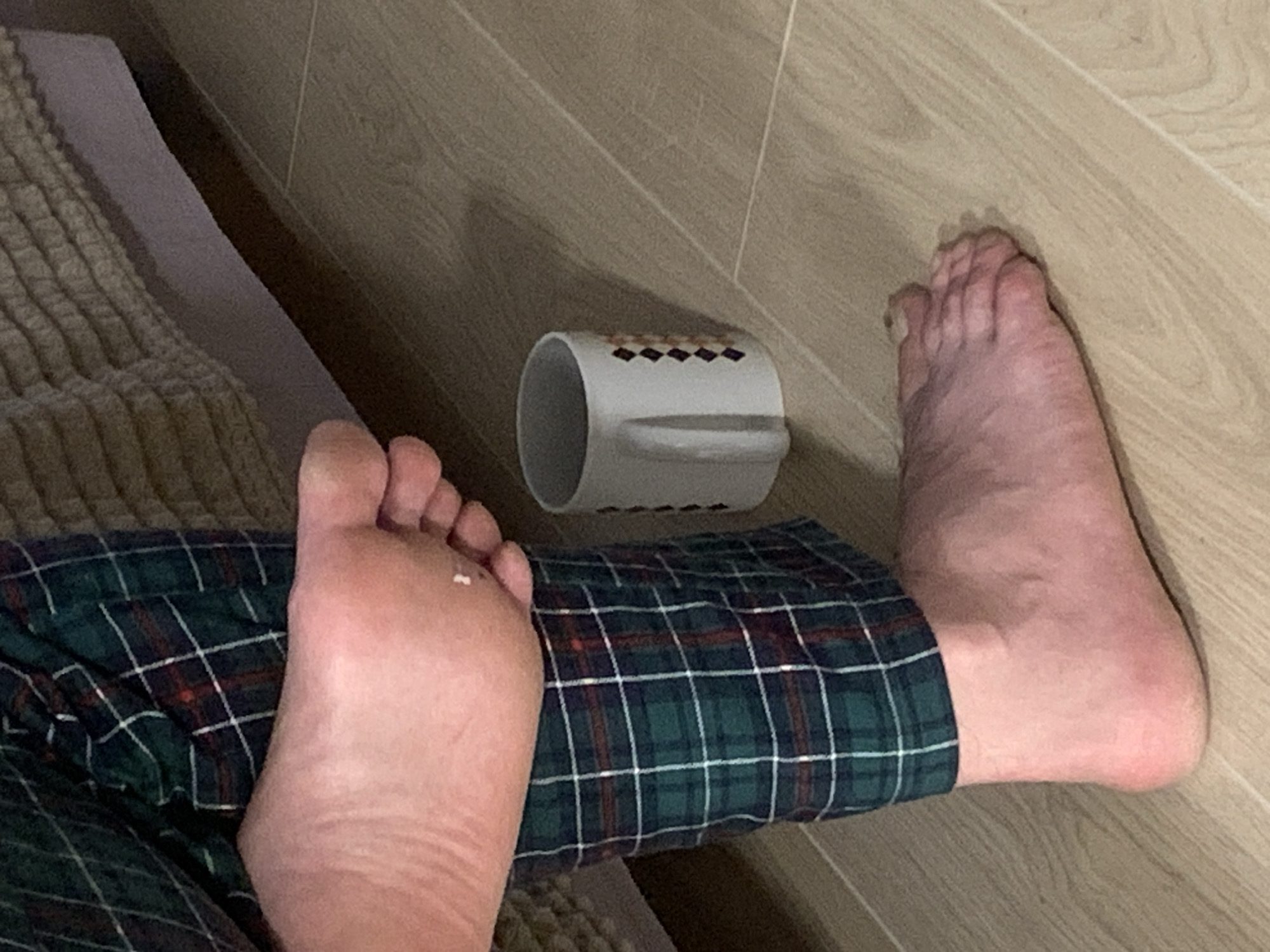 The image captures a close-up view of a person's feet and a white mug on a light-colored wooden floor. The person is wearing green and dark red plaid pajamas. One foot is resting on the other, revealing the sole of the upper foot. The mug, which appears to be ceramic, is positioned to the right of the feet. The background shows a textured surface, possibly a bed or sofa, and a hint of a wall. The scene is set indoors, with soft lighting. The location is identified as Kapyĺ, Belarus.