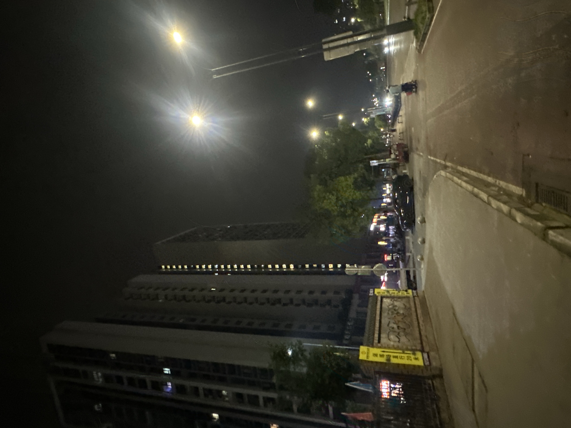 The image depicts a nighttime street scene in the Chongqing High-tech Zone, China. Tall apartment buildings line the left side of a paved road. Streetlights illuminate the scene, casting a glow on the wet pavement, suggesting recent rain. A few cars are parked along the sidewalk, and a person is visible riding a scooter or electric bike in the distance. The atmosphere is calm and quiet, with the urban environment lit by artificial lights.