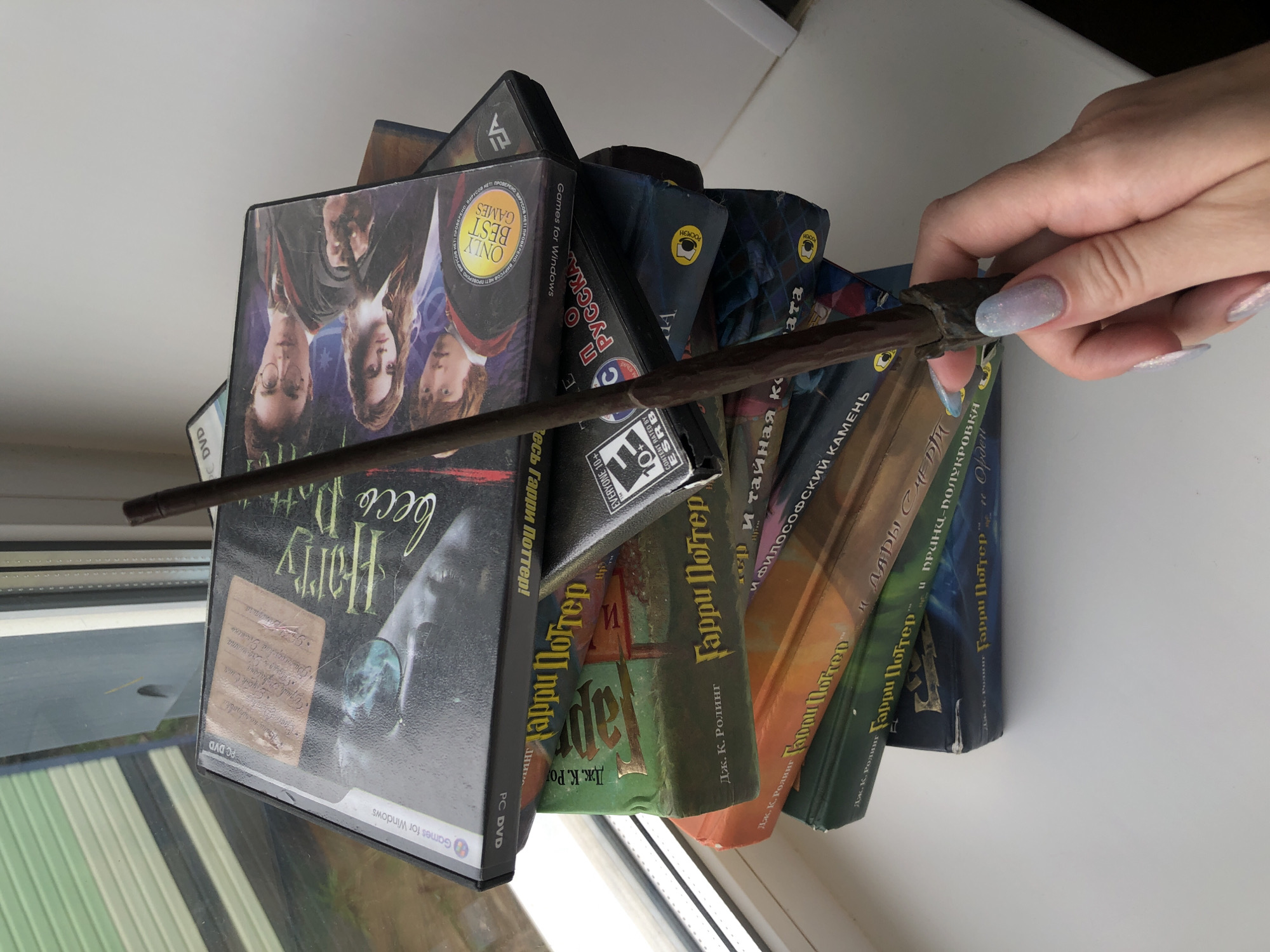 A hand is holding a Harry Potter wand over a stack of Harry Potter games and books in Russian. The books include: "Harry Potter and the Philosopher's Stone," "Harry Potter and the Deathly Hallows," "Harry Potter and the Half-Blood Prince," and "Harry Potter and the Order of the Phoenix." The location is a window in Noviy, Russia.