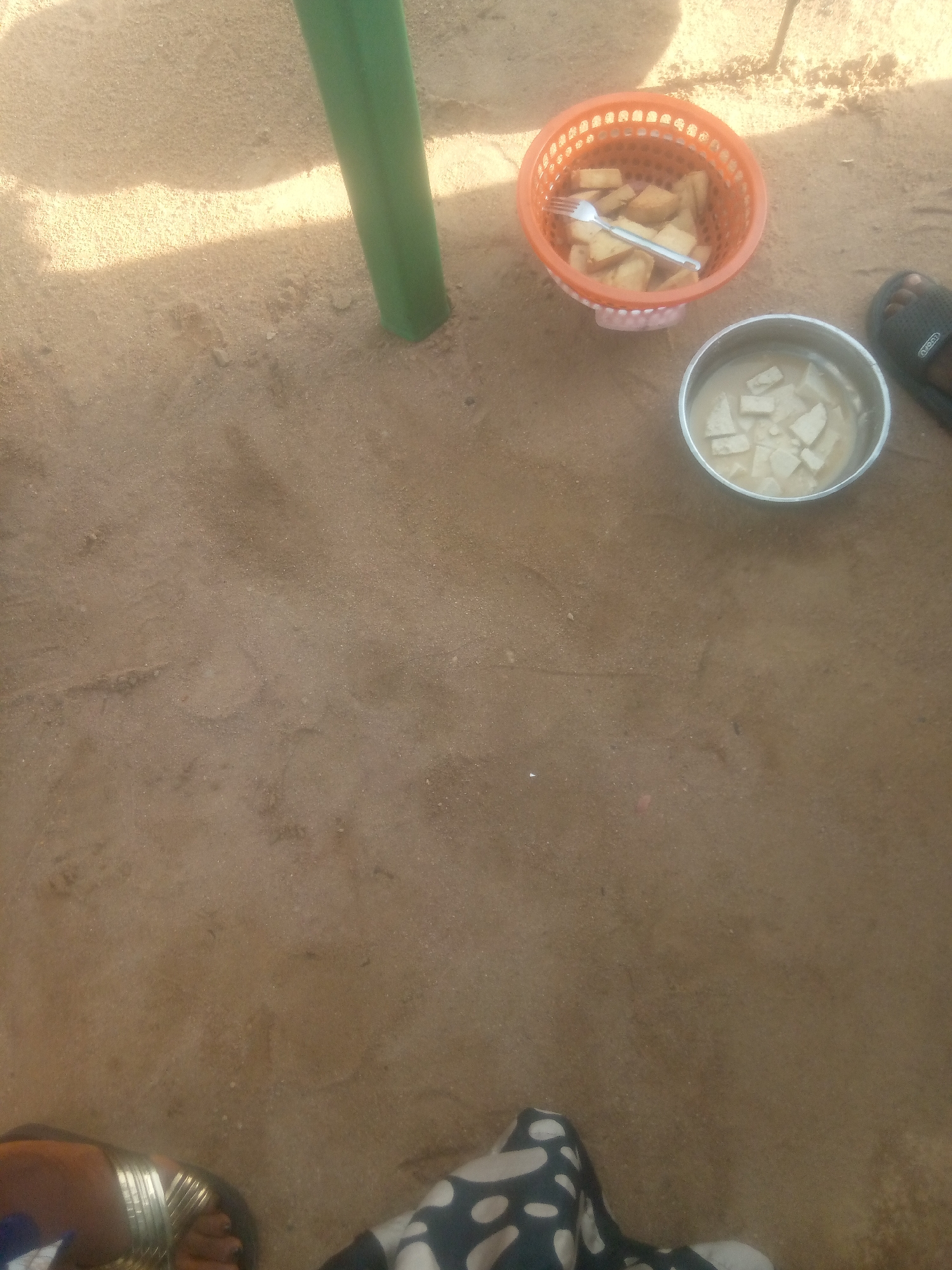 An overhead, high-angle shot shows a sandy ground with a green pole on the left side of the image. In the upper right quadrant, there is an orange plastic basket filled with cubed food and a fork. Next to the basket is a metal bowl containing a creamy liquid with cubed food. A black sandal is visible to the right of the bowl. The lower portion of the image shows the feet of two people. One person is wearing a sandal with a gold strap, and the other person is wearing patterned fabric. The scene is outdoors and appears to be in a casual setting.