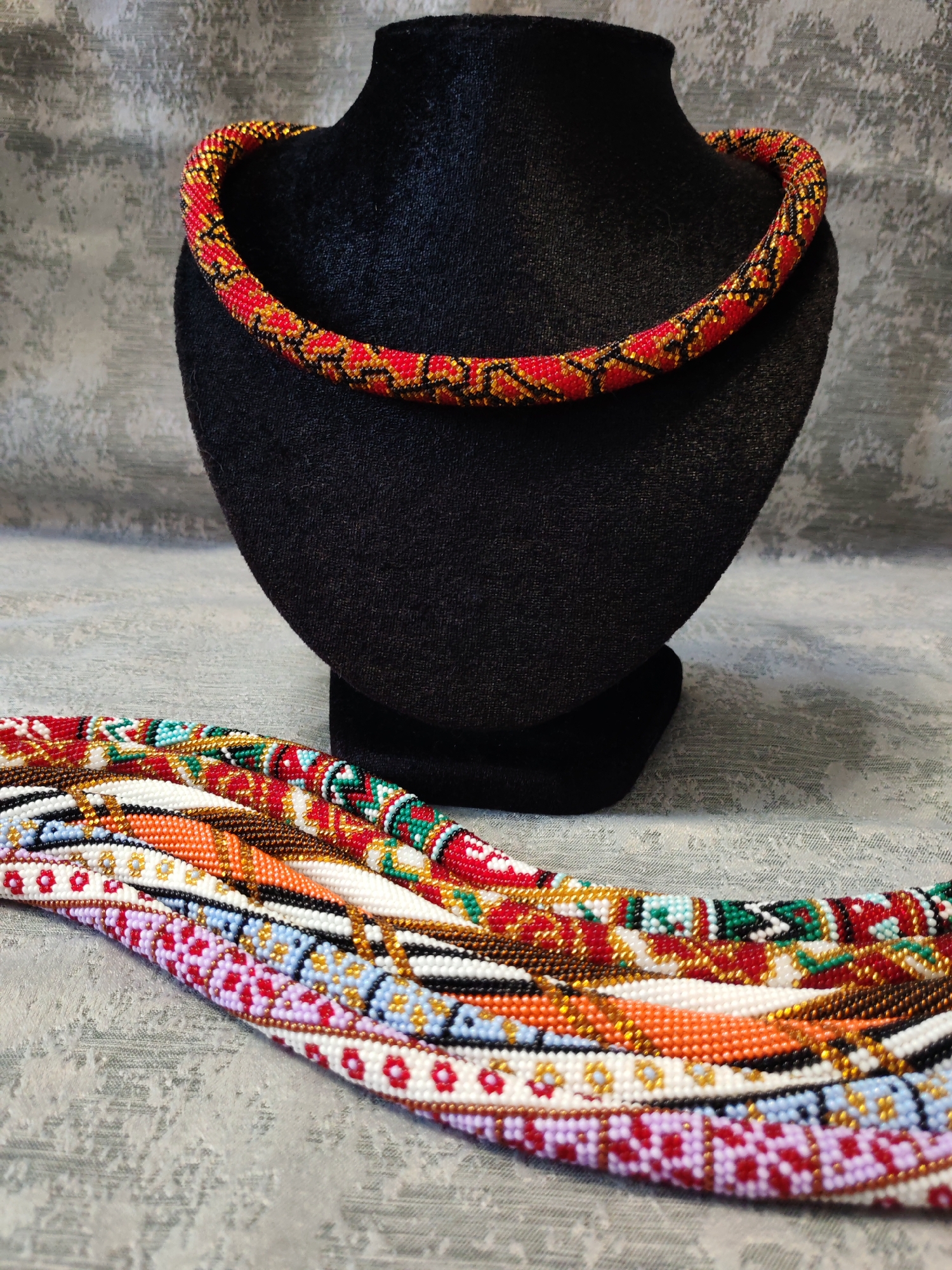The media file displays a collection of handcrafted beaded necklaces and a bracelet, arranged on a textured, light gray surface. In the background, a black velvet bust stands, showcasing a single, thick beaded necklace. This necklace is predominantly red, with intricate black and gold geometric patterns woven throughout its design.
In the foreground, several other beaded pieces are laid out. These appear to be a mix of necklaces and possibly a bracelet, all showcasing vibrant, multi-colored geometric patterns. The beads are small and tightly strung, forming rows and bands of color. Some of the visible patterns include designs with white, red, purple, light blue, orange, and green beads, interspersed with gold and black accents. The craftsmanship appears detailed and precise, with each piece exhibiting a distinct pattern and color scheme.
The lighting in the image is even, suggesting an indoor setting, likely a studio or a home environment where these handcrafted items are displayed or photographed. There are no people visible in the image, and no text is present. The focus is entirely on the artistry and detail of the beaded jewelry.