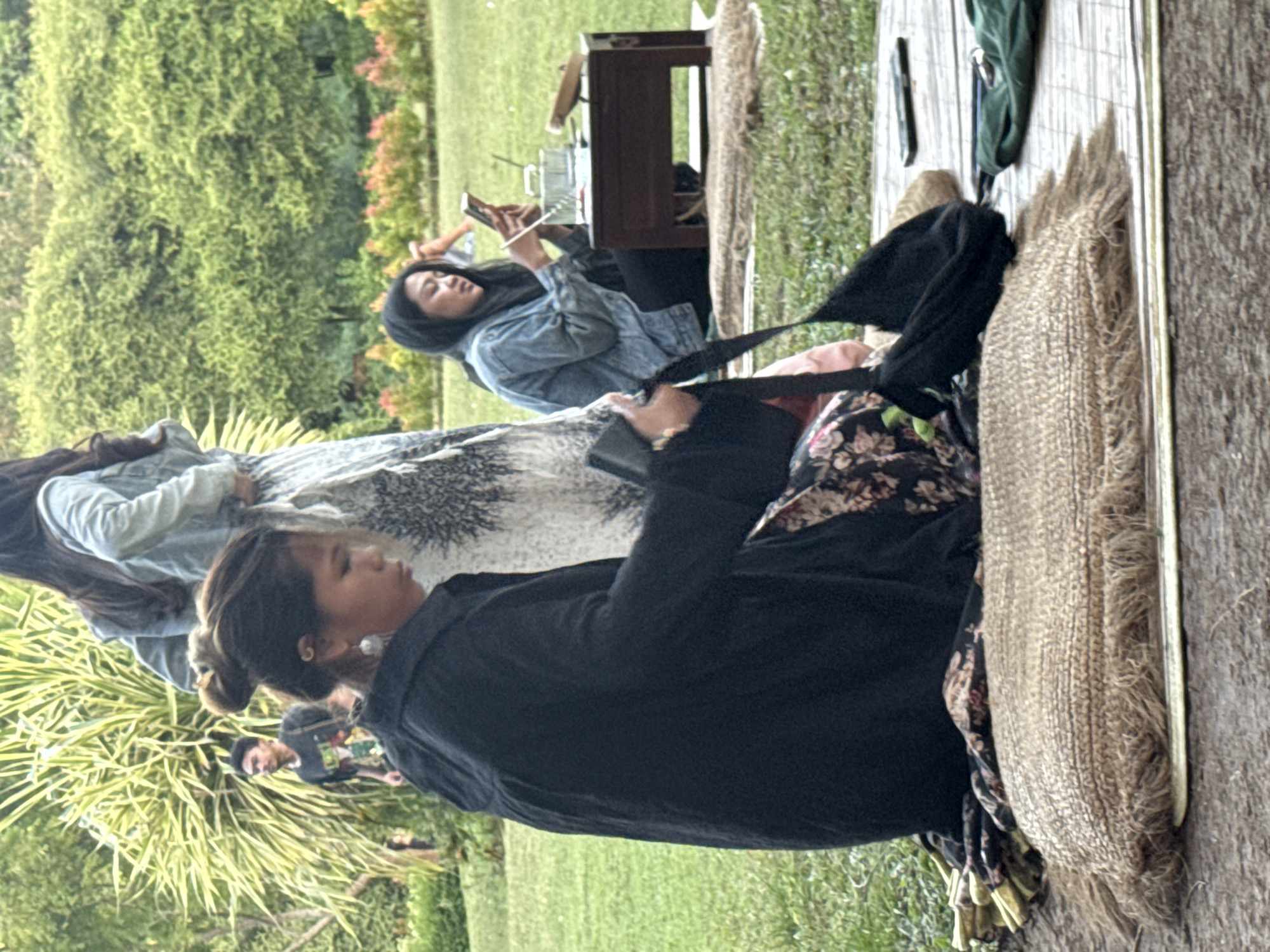 The image shows three women sitting outdoors on a wooden platform, possibly in Pecatu, Indonesia, given the context. The woman closest to the camera is wearing a black top and floral print pants. She's positioned on a fringed mat and appears to be adjusting a black strap. A second woman, wearing a denim jacket, is seated behind her and looking at a mobile phone. A wooden table and some greenery are also visible. The background includes a grassy area and dense trees. The scene suggests a relaxed, casual atmosphere.