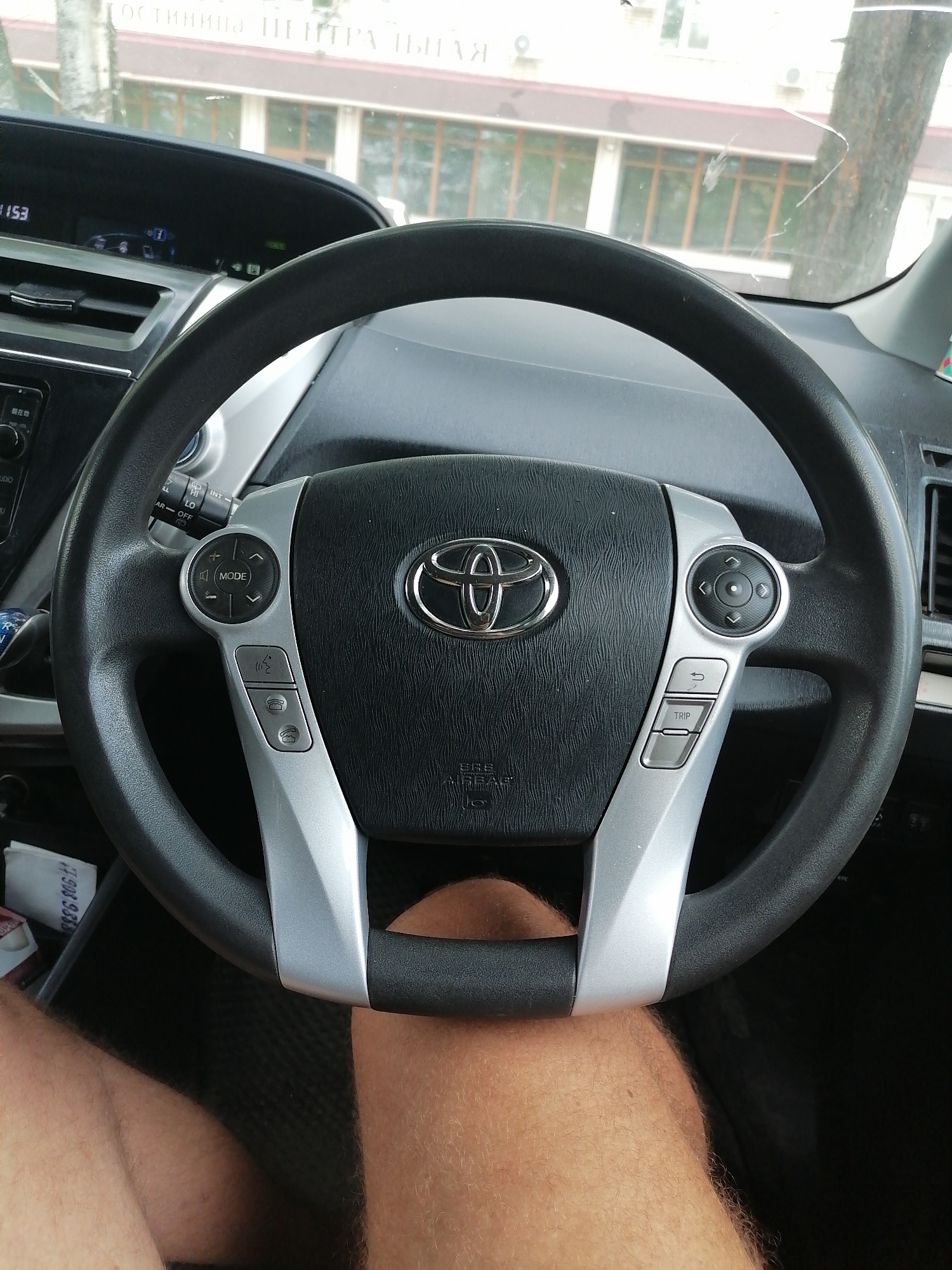 A close-up, eye-level shot shows the interior of a car, specifically the steering wheel and dashboard, from the driver's perspective. The steering wheel, centrally located, features the Toyota logo and various control buttons. The dashboard displays a digital clock reading "1153" and other indicators. Outside the car, through the windshield, a building with multiple windows and a tree are visible. The image appears to be taken in the city of Lesozavodsk, Russia, based on contextual information not present in the image itself.