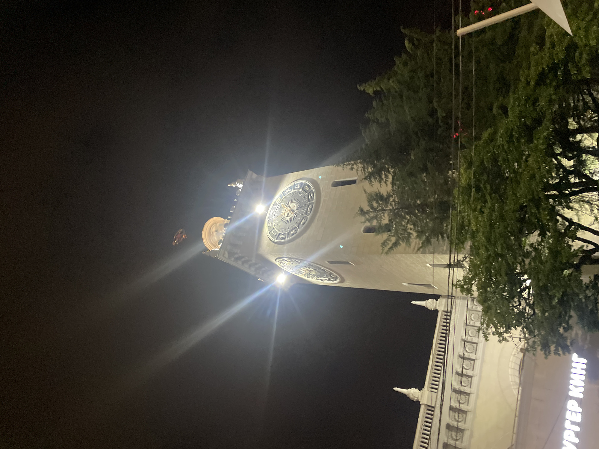 The image is a nighttime, low-angle shot of a tower with two large, ornate clocks on it. The tower is made of light-colored stone and has architectural details on its upper section. A bright light shines from the top of the tower, causing lens flare in the image. The tower is surrounded by dark trees. In the lower left of the image, a sign with white illuminated letters, partially visible, reads "УРГЕР КИНГ" which likely translates to "BURGER KING". The overall scene suggests an urban setting in Sochi, Russia, possibly a public square or a prominent building. The sky is dark, indicating it is nighttime.
