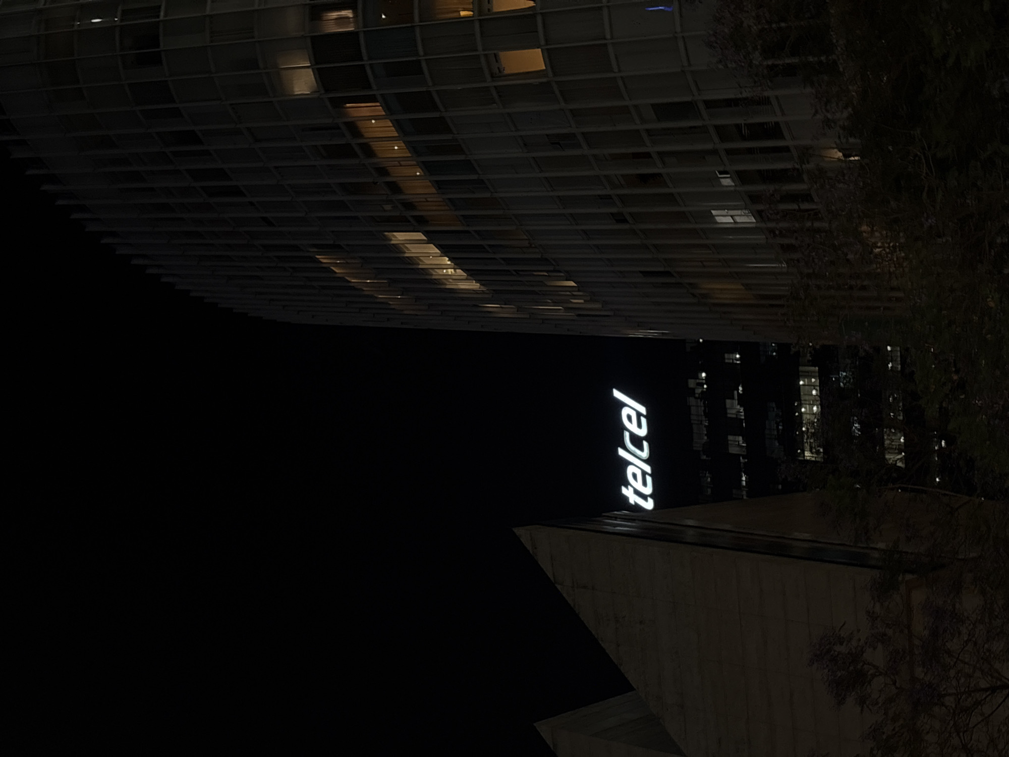 Captured at night in Mexico City, Mexico, the image presents an unusual perspective of the Telcel building. The illuminated "telcel" logo is prominently displayed against the dark sky. The building has a distinct architectural structure, possibly a monument or a corporate building with modern design elements, visible in the lower right and center. Above the building is another, much taller and wider, building with an exoskeleton or grid-like facade. This building is lit from within in a few places. To the right are the hazy lights of buildings further away, and the blurred silhouette of tree branches. The environment appears urban, characterized by tall buildings. The overall scene is lit by the glow of the Telcel sign and some light from the buildings.