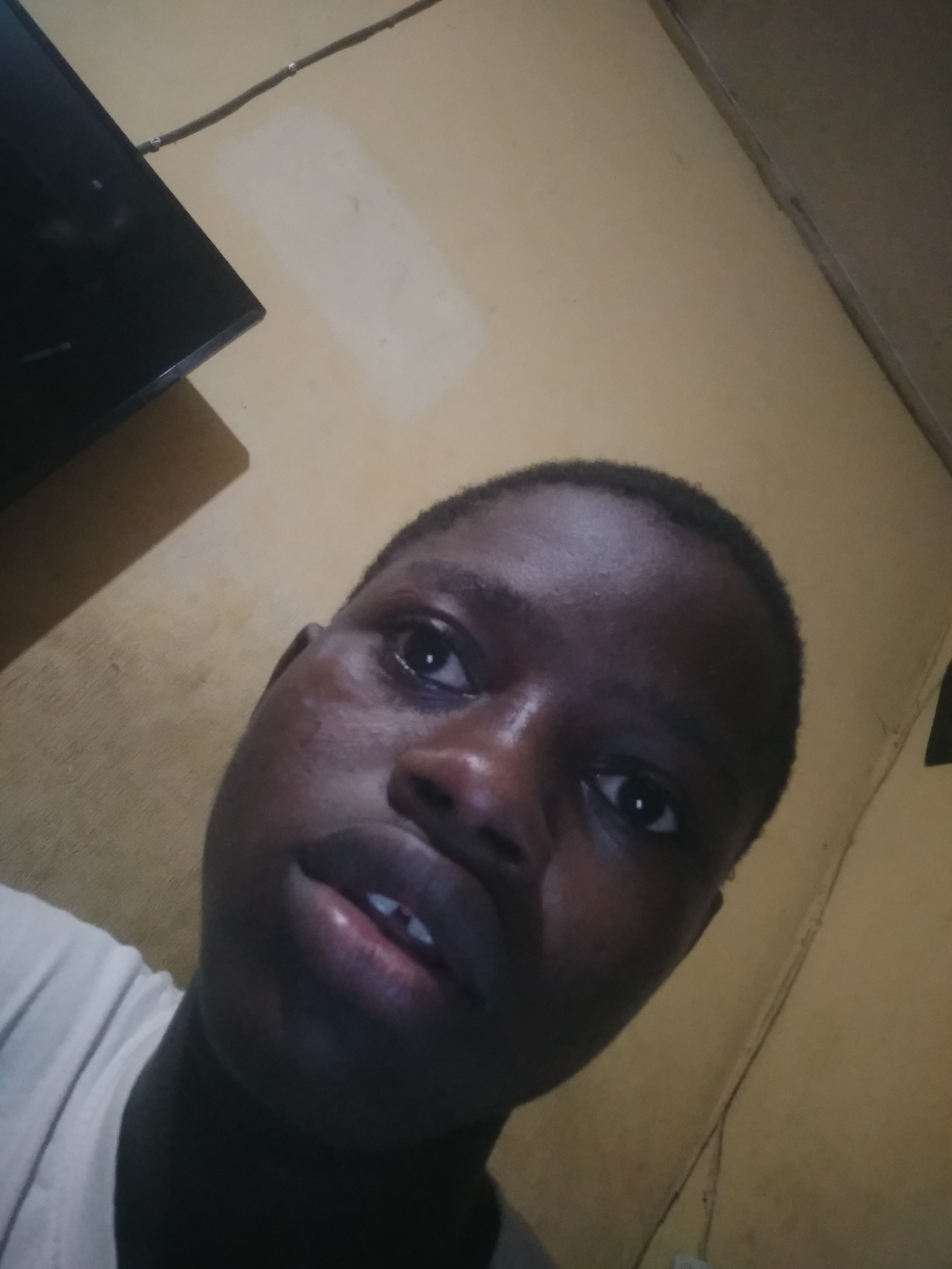 A close-up, low-angle selfie of a young person with dark skin and short hair. The individual's face is the primary subject, featuring dark eyes, a wide nose, and full lips. The person is wearing a white shirt or t-shirt. The background includes a portion of a dark-colored television screen in the upper left corner and a light yellow wall. The location is Unknown, Unknown.