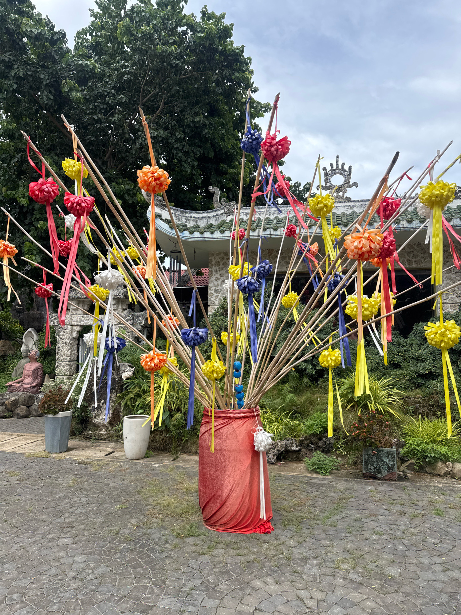 The image shows an outdoor scene in Tuy An Nam, Vietnam, featuring a vibrant display of decorative elements. A large arrangement of bamboo poles stands prominently in the foreground, adorned with numerous colorful paper flowers and ribbons in red, yellow, orange, blue, and white. These decorations are suspended at varying heights, creating a festive and dynamic visual. The bamboo poles are bound together at their base, which is wrapped in a red cloth.
Behind the display, traditional Vietnamese architecture is visible, characterized by a curved, tiled roof and ornate decorative elements on the roofline. The building appears to be part of a cultural or religious site. Lush greenery surrounds the structures, with trees and various plants forming a dense backdrop.
In the left midground, a stone statue of a seated figure, possibly a monk, rests on a pile of rocks. Several large planters, one gray and one white, are positioned near the statue and the main decorative arrangement. The ground is paved with interlocking stone tiles, forming a circular pattern. The sky above is overcast with clouds, suggesting a diffused light condition. The overall scene conveys a sense of cultural celebration or spiritual observance.