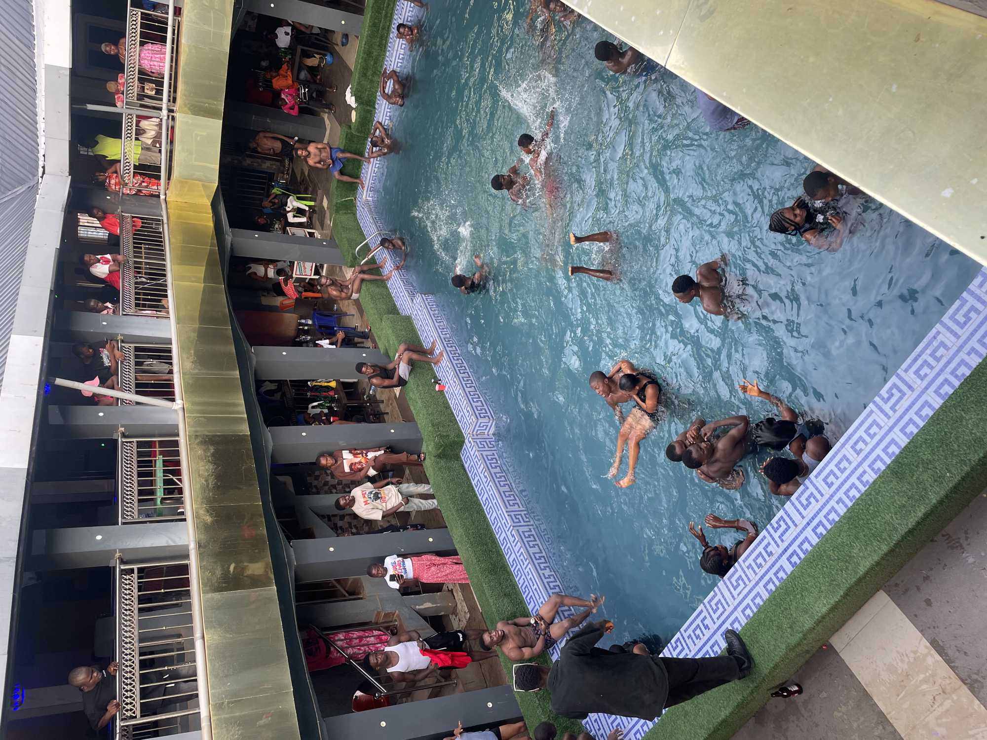 The image shows an outdoor swimming pool at a building with multiple balconies in Onitsha, Nigeria. Numerous people are in the pool, swimming, splashing, and interacting with each other. Several other people are either sitting or standing along the pool's edge. The building surrounding the pool features balconies with people observing the scene. The pool has a blue and white tiled border and is surrounded by artificial turf. The building has a gold-colored facade and grey balconies. Some people are standing on the balconies.