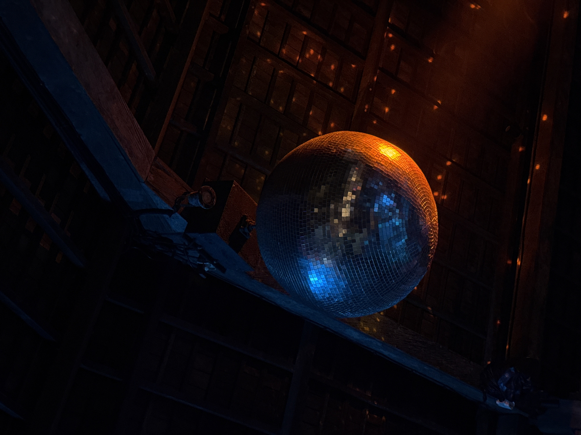 A low-angle shot reveals a disco ball suspended from the ceiling in a dimly lit space. The disco ball's reflective tiles create dynamic patterns of light and shadow. The surrounding environment consists of a dark ceiling structure with wooden beams. Some of the light glints off the ceiling creating scattered small orange spots. The photo may have been taken in North Kuta, Indonesia.