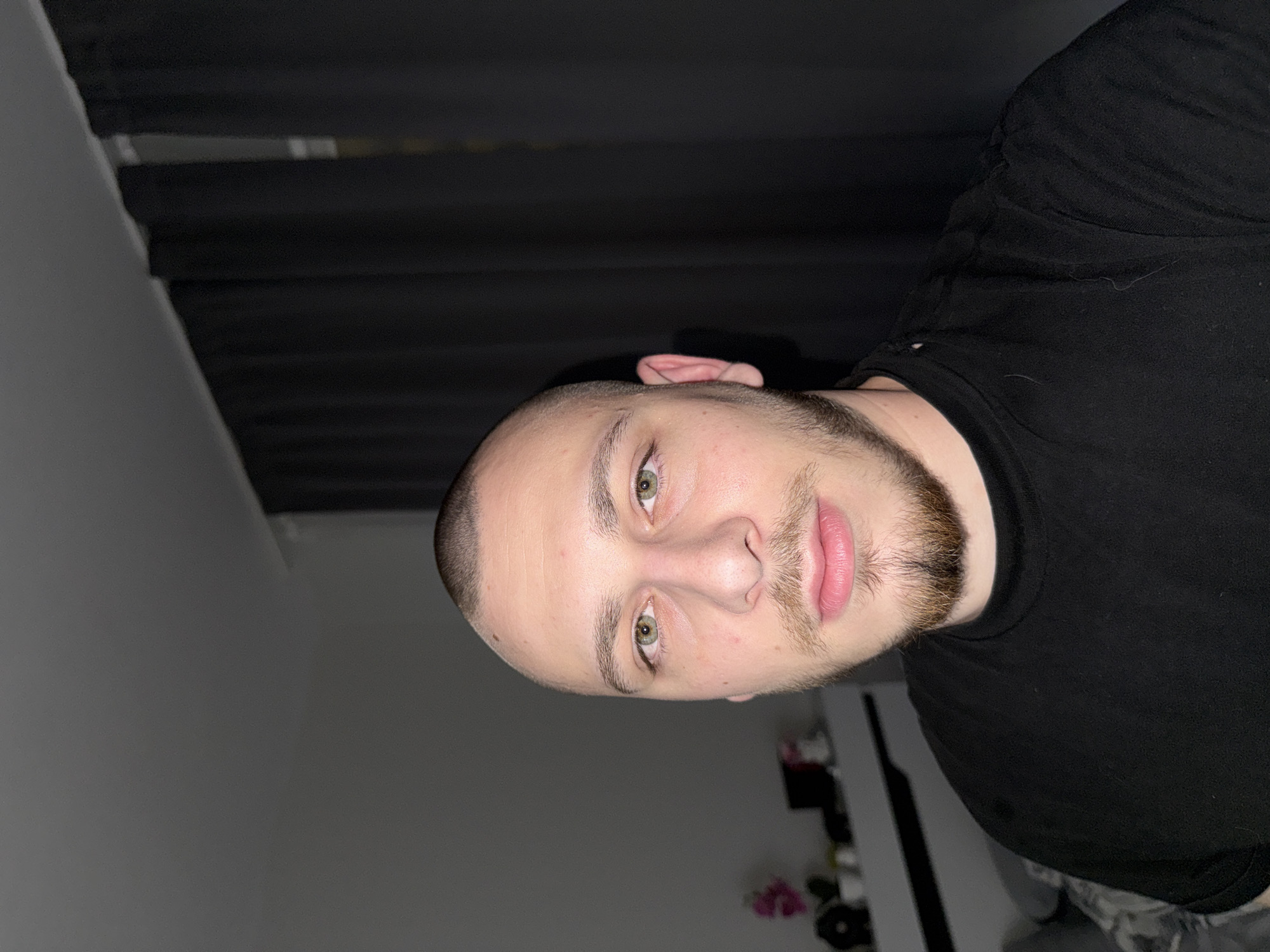 A medium close-up, portrait-style photograph shows a fair-skinned male with light brown hair shaved very close to the scalp. He has green eyes and a light brown beard. He is wearing a black t-shirt. The background is a dimly lit room with a partially obscured window with a dark curtain. The location is impossible to determine based solely on the image.