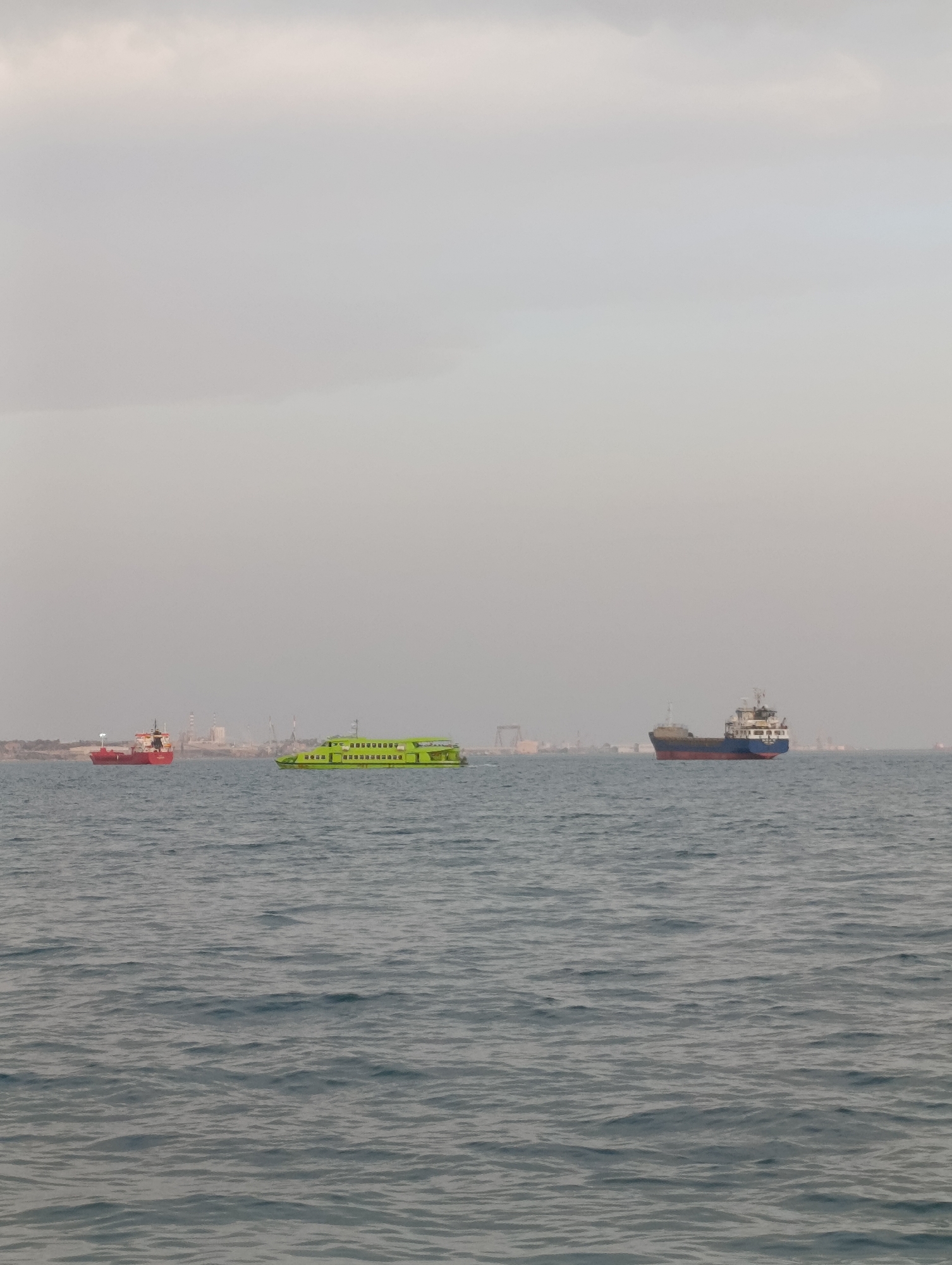 An overcast sky looms over a body of water with three vessels. On the left, a red cargo ship is positioned near a hazy coastline with industrial structures. In the center, a bright green passenger ferry moves across the water. To the right, a blue and brown cargo ship is visible on the horizon. The scene is likely Setúbal, Portugal, given the industrial architecture and presence of ferries.