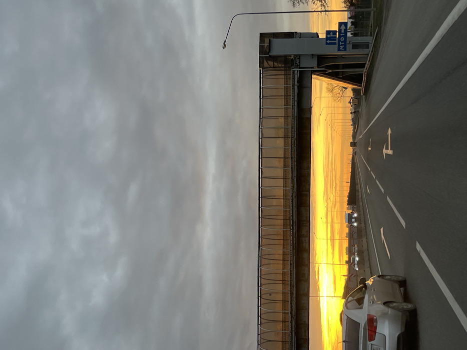 The image depicts a multi-lane highway at either sunrise or sunset, situated in the city of Lenina, Russia. The sky overhead is heavily overcast with grey clouds, transitioning to a brilliant orange and yellow hue along the horizon, which casts a warm glow across the scene.
In the foreground, a silver four-door sedan is visible on the left side of the road, traveling away from the viewer. The asphalt road surface features white dashed lane markings and prominent white arrows indicating both straight-ahead and right-turn options in the lanes.
Dominating the midground is an elevated pedestrian bridge spanning the entire width of the highway. Its enclosed walkway is constructed with translucent, amber-toned panels that intensely reflect the golden light from the horizon. The bridge's concrete and metal supports are visible on the right. Below the bridge, on the right shoulder, a blue road sign displays Cyrillic text "МТФ1" with an arrow indicating both straight and right directions.
Numerous streetlights line both sides of the highway, extending into the distant background. Several other vehicles, including cars and a larger white truck or van with illuminated headlights, are visible further down the road, also traveling away from the camera. The distant horizon reveals faint silhouettes of urban structures against the bright sky, and a few bare trees are discernible on the right side of the highway. No individuals are explicitly visible in the scene.
