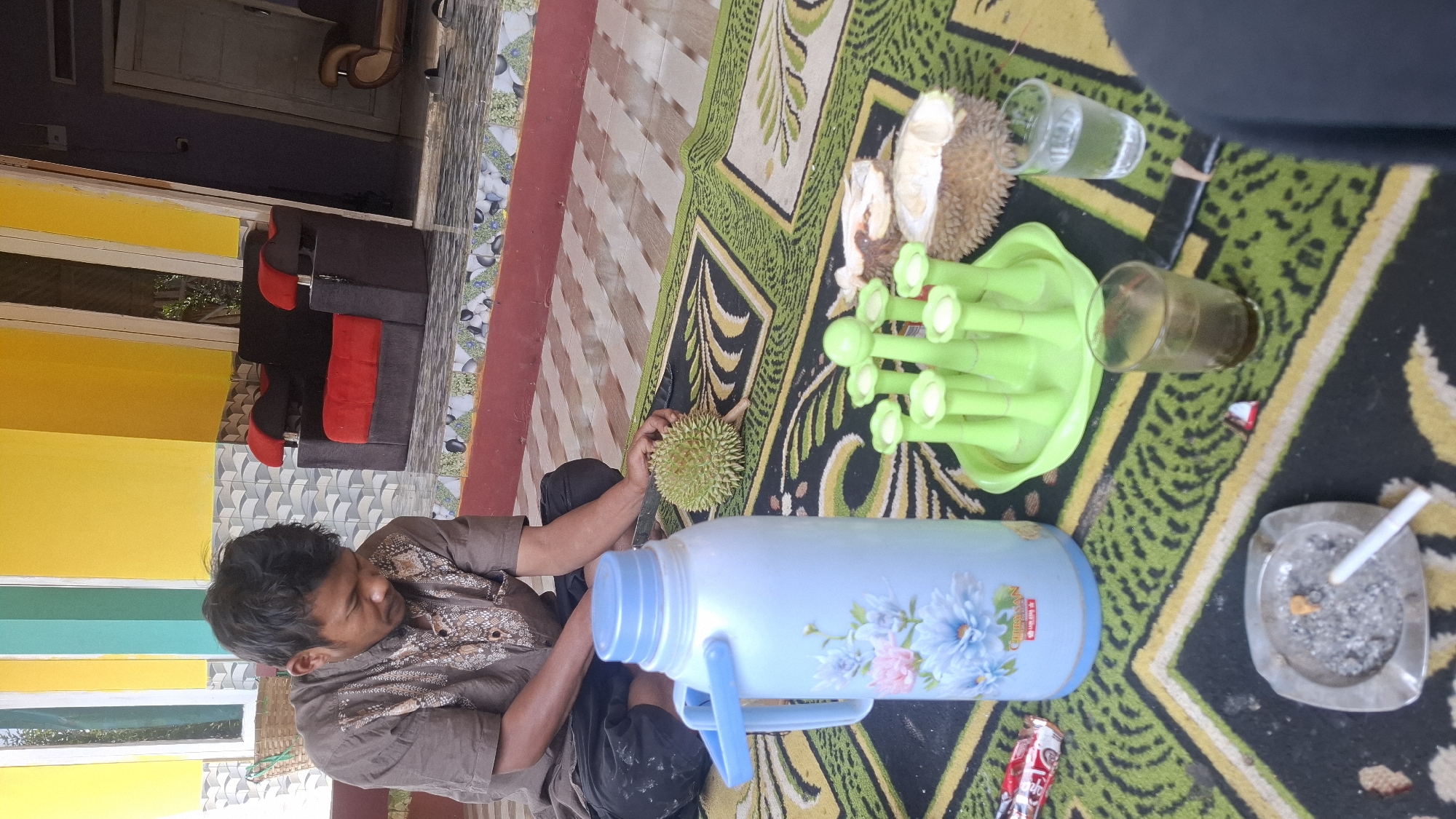 In Cilaku, Indonesia, a man sits cross-legged on a patterned rug, preparing to cut a durian fruit. He is dressed in a brown, patterned shirt and dark pants, with a knife in his right hand, poised above the spiky fruit. Nearby, a light blue thermos with a floral design stands upright. A green plastic tray holds several green, stem-like objects, possibly for serving. Two pieces of durian, one cut open to reveal its flesh, are placed on the rug. Two glasses, one containing a dark liquid and the other clear water, are also present. An ashtray with cigarette butts and ashes sits on the rug, with a lit cigarette resting in it. In the background, a red and black sofa is visible, as is a yellow wall and patterned tiles. The scene appears to be outdoors, possibly on a porch or patio.