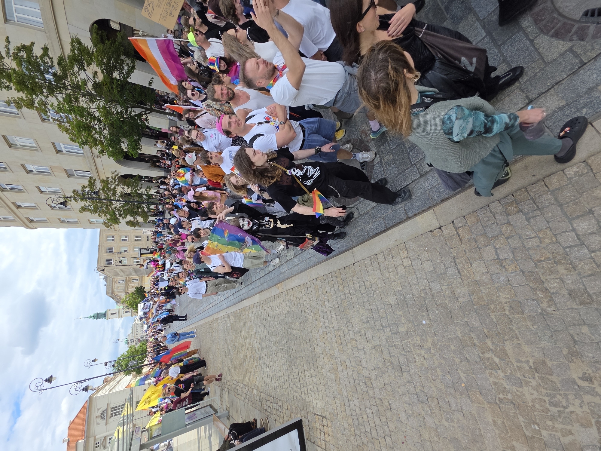 The image depicts a vibrant street parade in Warsaw, Poland, likely a Pride event, given the presence of rainbow flags and symbolic attire. A diverse crowd of people walks along a cobblestone street lined with buildings. In the background, a green spire of a church is visible, along with a light-colored, multi-story building with arched entrances. Several street lamps with ornate fixtures stand along the parade route. People are dressed in a variety of casual clothing, with some holding rainbow flags, banners, and balloons. One individual in the foreground wears face paint and a skeleton-themed outfit. The atmosphere appears festive and celebratory, with participants engaging with each other and some capturing the event with their phones. The street is paved with light-colored cobblestones, and a concrete sidewalk runs alongside the buildings.