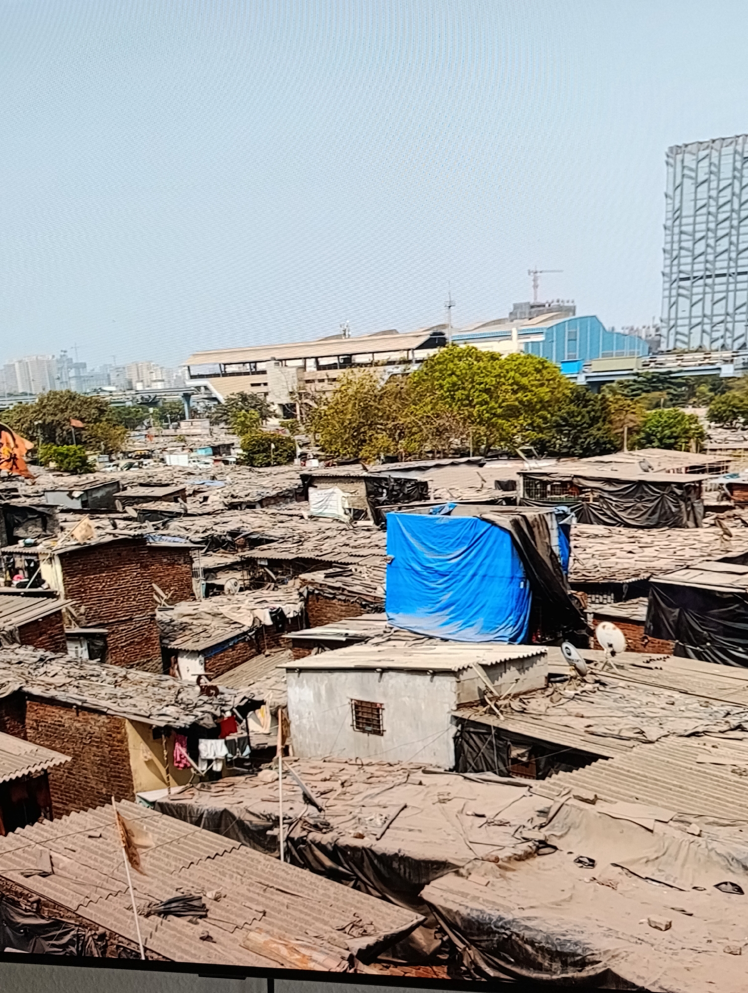 The image shows an aerial view of a densely populated slum in an unknown city. The foreground is dominated by closely packed makeshift dwellings with corrugated iron or tarp roofs. Some roofs are covered with black plastic sheeting, while others show signs of age and decay.
Several structures appear to be built of brick. Clothes are hanging out to dry on a line strung between buildings. A bright blue tarp covers one of the structures. A satellite dish is visible on a rooftop.
In the background, a modern building is under construction, suggesting a juxtaposition of poverty and development. A transportation hub or station with a blue roof is also visible, and a few high-rise buildings are silhouetted against the clear sky. Trees and sparse greenery are scattered throughout the scene.