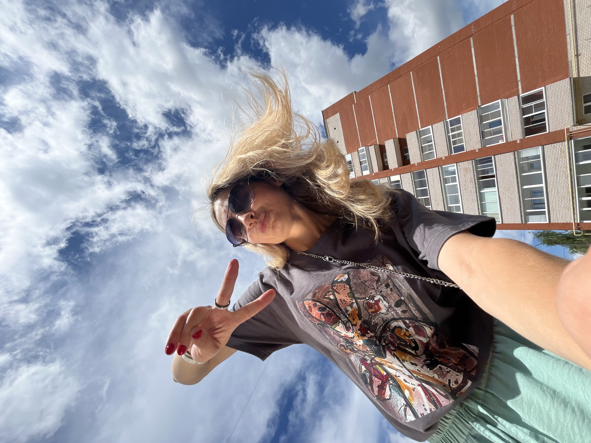 An outdoor, low-angle, eye-level, medium shot shows a blonde woman in sunglasses and a dark grey t-shirt with a graphic print and a light green skirt, posing for a selfie. Her hair is blowing in the wind. She is making a peace sign with her right hand, and her lips are puckered. Behind her, a tall, red brick apartment building with multiple windows and balconies dominates the background, set against a bright blue sky with scattered white clouds. The image appears to have been taken in the city of Chaikovsky, Russia, based on the architecture of the building and the surrounding environment which is consistent with residential areas in such cities.