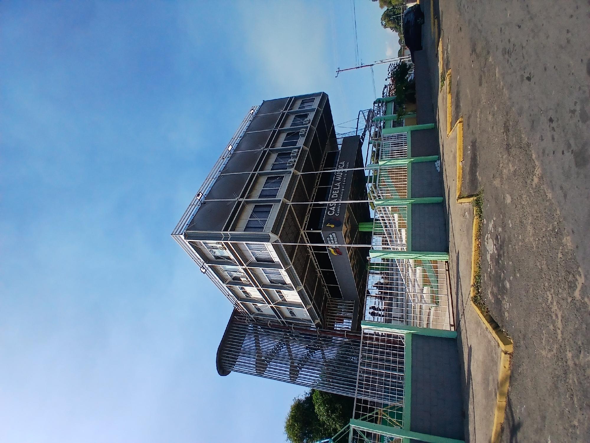 The image shows the "Casa de la Musica" (House of Music) building in Ciudad Guayana, Venezuela. The building is a multi-story structure with a modern design, featuring a grid-like facade. The building is surrounded by a green and white metal fence and a sidewalk. A road with yellow painted edges is visible in the foreground. The sky is blue with some wisps of clouds. A few trees and a dark-colored vehicle are visible in the background. The image is taken from a low angle, looking up at the building.