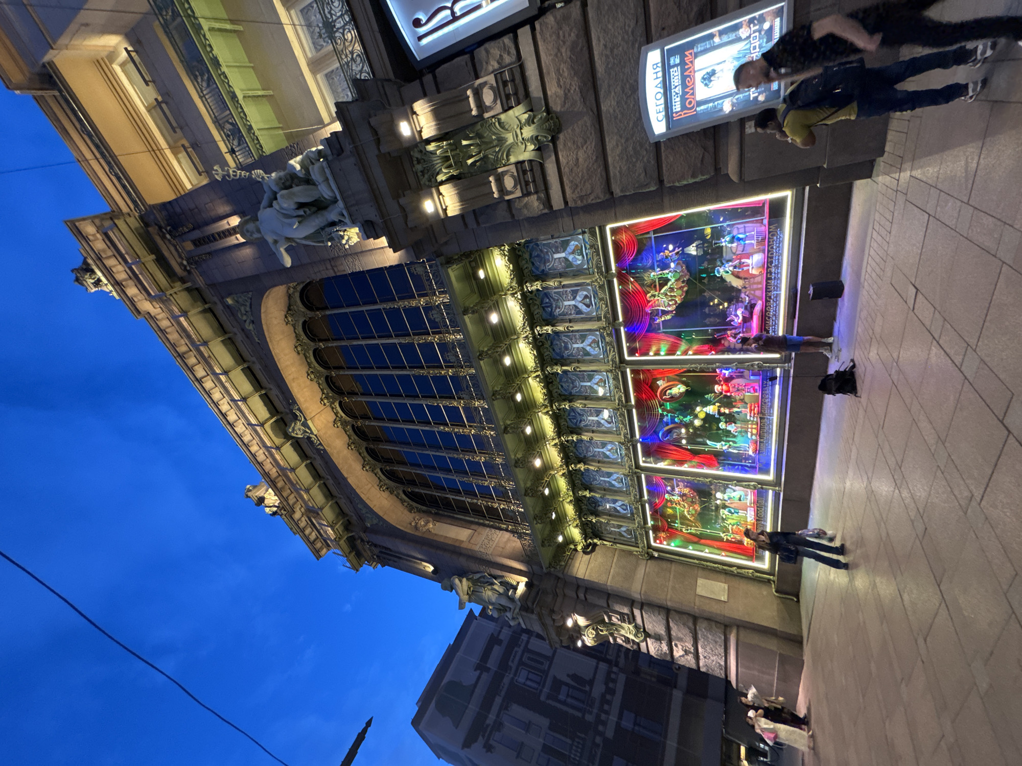 The image captures the Singer House, also known as the Dom Knigi, on Nevsky Prospekt in Saint Petersburg, Russia, during the evening. The grand Art Nouveau building is illuminated, highlighting its elaborate facade. The large, arched windows of the Singer House are adorned with decorative elements and bright lights. Below these, the ground floor features display windows showcasing colorful and vibrant scenes, likely for a holiday or event, with red curtains framing the exhibits.
Several people are visible on the sidewalk in front of the building. On the left, a group of women are walking, one in a white dress. To the right of the display windows, a person stands looking at the exhibition. Further to the right, two men are walking, one wearing a yellow shirt and backpack, the other in a dark patterned shirt. A third person is walking away from the camera in the foreground, carrying a bag. A portable speaker sits on the ground near one of the display windows. A large illuminated poster board stands on the right side of the building, displaying text and an image. The sky is a deep blue, indicating it is dusk or early evening. The cobblestone sidewalk and the architectural details of the building are clearly visible.