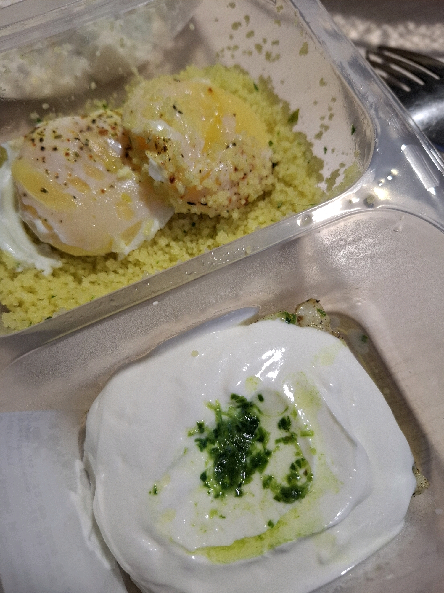 A close-up, top-down view captures two servings of food in a clear plastic container. The upper serving consists of fluffy yellow couscous topped with two poached eggs, seasoned with herbs and spices. The lower serving is a generous dollop of white yogurt or cheese, garnished with chopped green herbs and a drizzle of oil. The container appears to be takeaway packaging. The background is blurred, showing a neutral surface, possibly a table, and a hint of cutlery. No people are visible in the image. The scene is indoors, likely a restaurant or home setting. There is no indication of the city of Tyumen, Russia.