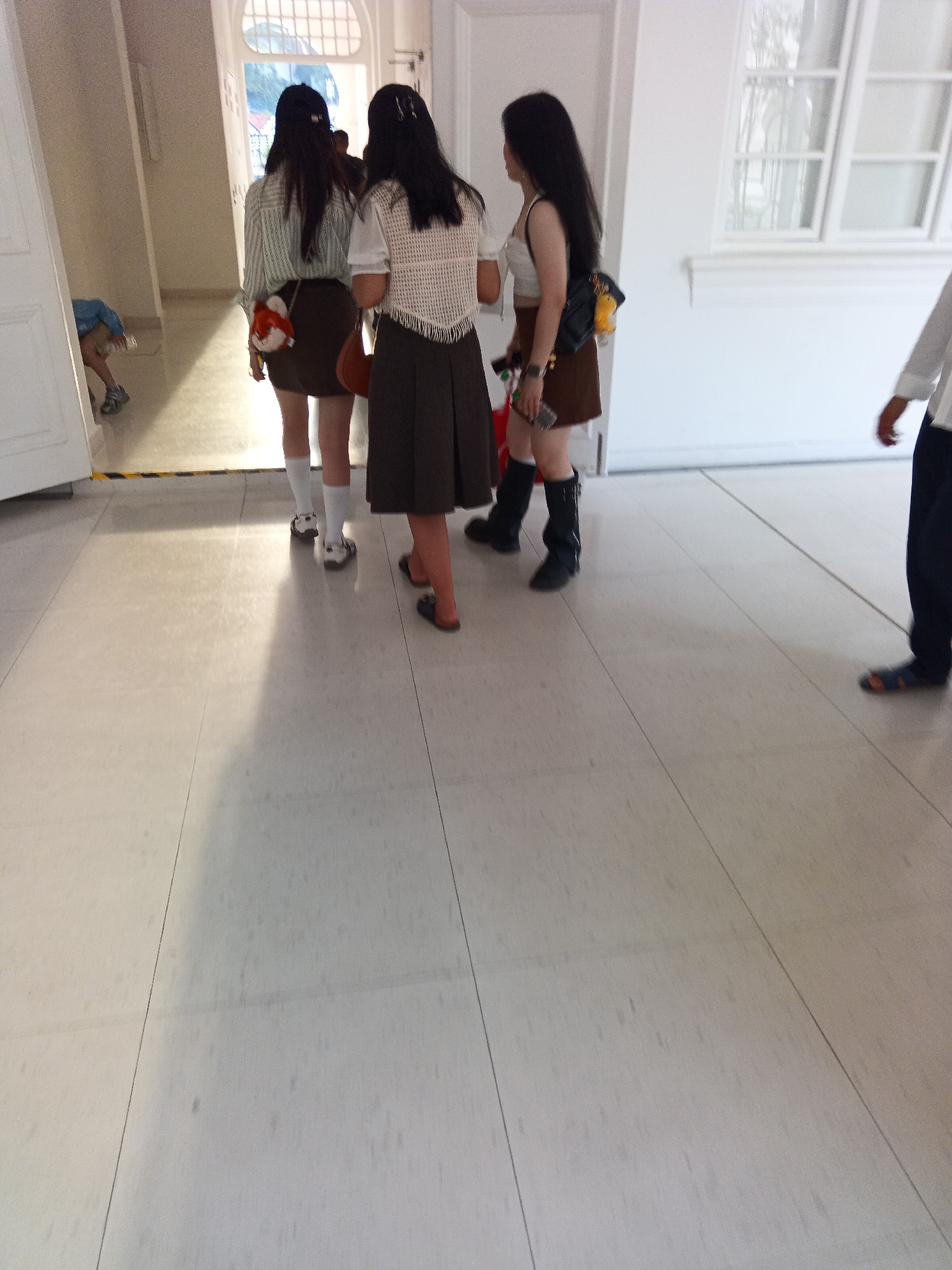 Three young women, appearing to be in their late teens or early twenties, are standing in a hallway with tiled flooring. The woman on the left wears a vertically striped long-sleeved shirt, a dark brown skirt, white socks, and white sneakers. She has a small orange bag hanging from her shoulder. The woman in the middle is dressed in a white crocheted top with a fringed hem, a dark brown skirt, white socks, and white sneakers. She is holding a smartphone. The woman on the right wears a white tank top, a dark brown skirt, and black knee-high boots. She is carrying a dark bag with a yellow charm and a brown bag over her shoulder. A fourth person, an adult male, is partially visible on the right side of the image, wearing dark pants and blue sandals. The hallway features a large window with mullions on the right and a doorway on the left. The overall impression is that of a casual gathering or a public space like a school or an institution. The scene does not provide specific indicators for a location within Kuala Lumpur, Malaysia, beyond being an indoor environment.