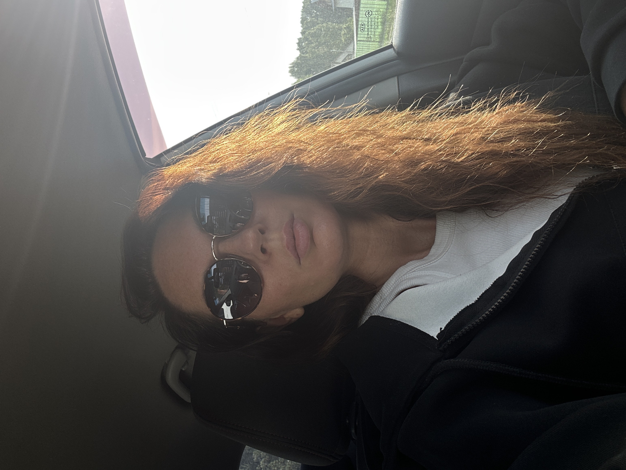 A young woman with long, reddish-brown hair, wearing sunglasses and a black zip-up hoodie over a white t-shirt, is pictured inside a car. She is looking directly at the camera. The sun is shining brightly, casting a warm glow on her hair. The interior of the car is visible, including a headrest and part of the dashboard. Through the car window, a blurry background of trees and a building can be seen, suggesting an outdoor setting. The car appears to be parked on a street in Chaikovsky, Russia.