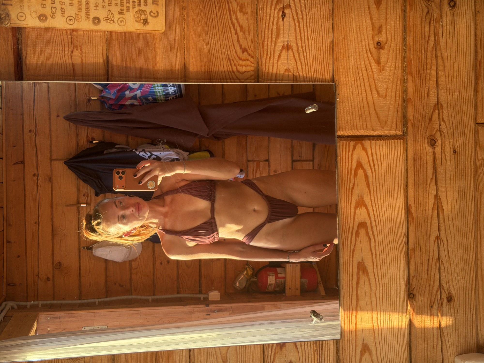 A woman with blonde hair pulled into a ponytail, wearing a burgundy bikini, stands in front of a wooden-paneled wall and takes a mirror selfie. She is holding a phone in her right hand. Behind her, several items are hanging on hooks, including clothing and a white cap. A fire extinguisher is visible on a shelf to her left. A sign with text and drawings is mounted on the wall to her right. The scene appears to be indoors, possibly in a rustic cabin or sauna. The lighting suggests it is daytime, with sunlight casting shadows on the wooden surfaces.