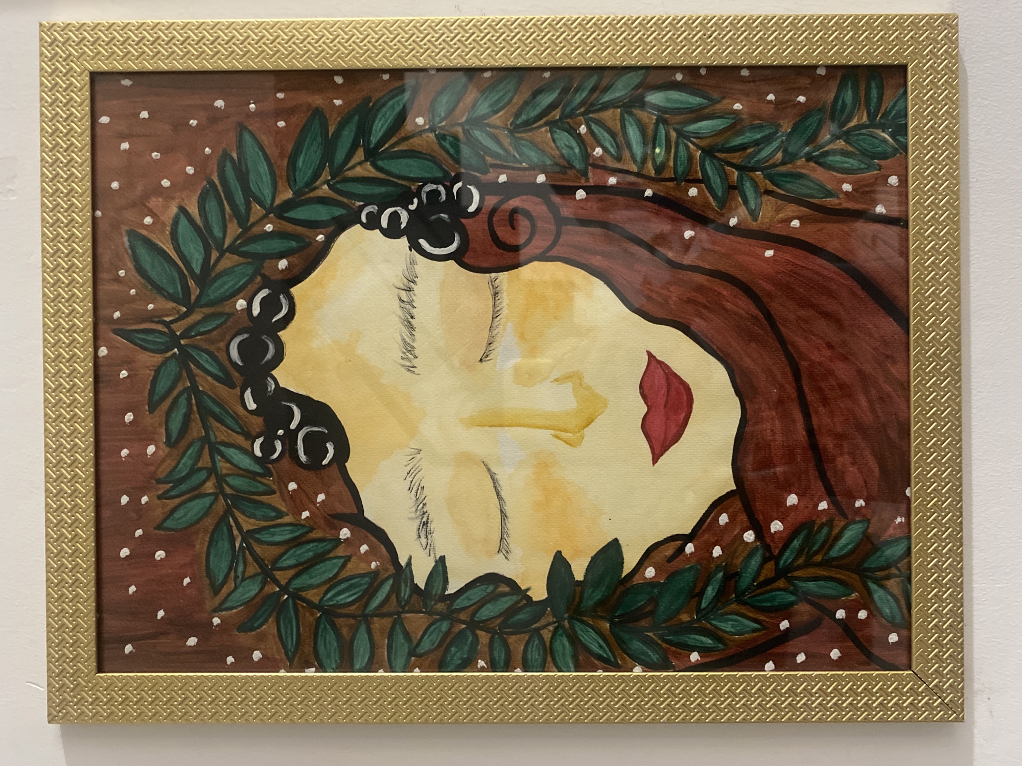 A framed painting of a woman with closed eyes, surrounded by a wreath of green leaves, hangs on a white wall. The woman has brown hair and red lips. The background is brown with white dots. The frame is gold with a textured pattern. The location is not discernable from the image.