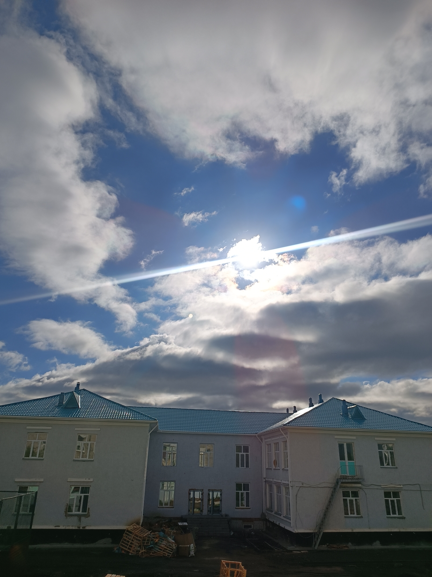The image shows a two-story building with a light blue roof under a partly cloudy sky, possibly located in Kokshetau, Kazakhstan. The building has multiple windows and an external staircase leading to a door on the second floor of one section. The building's exterior is painted in a light grey. Some wooden pallets and debris are on the ground in front of the building. The sky is blue with scattered white and grey clouds, and the sun is visible, creating a lens flare effect in the image. The ground in front of the building appears to be dark and possibly muddy.