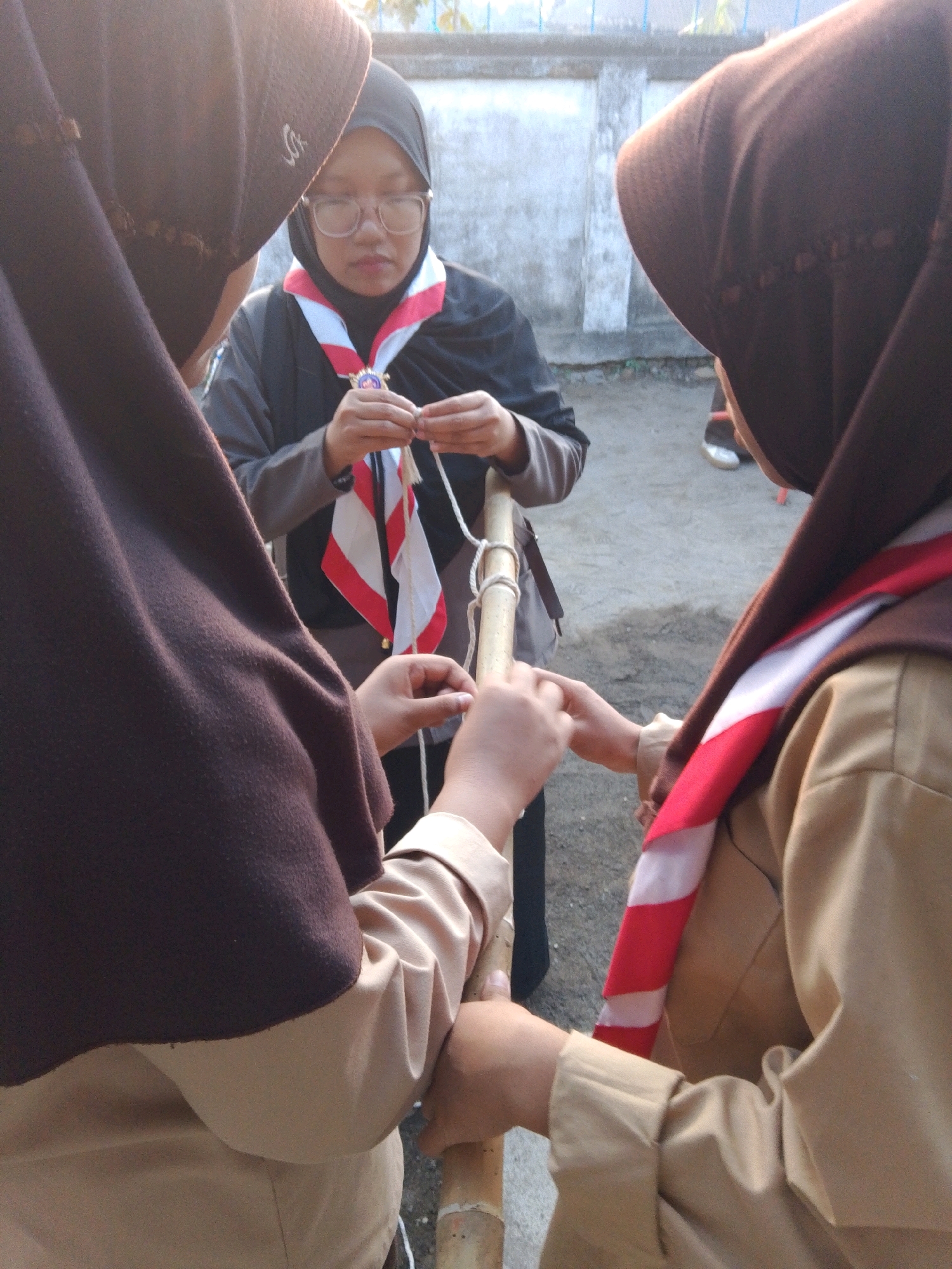 In a low-angle outdoor shot, three Indonesian Scout members in uniform are engaged in a knot-tying activity with a wooden pole. The Scout on the far left, facing away from the camera, wears a dark brown hijab and tan uniform shirt. To her right, a Scout wearing glasses, a black hijab, and a tan uniform shirt, is focused on manipulating a white rope with red and white striped neckerchief draped around her neck. On the right, another Scout, partially visible and facing the pole, also wears a tan uniform and brown hijab, with her hands assisting in the knot. The background features a gray concrete wall and a dirt ground, suggesting a simple outdoor training area.