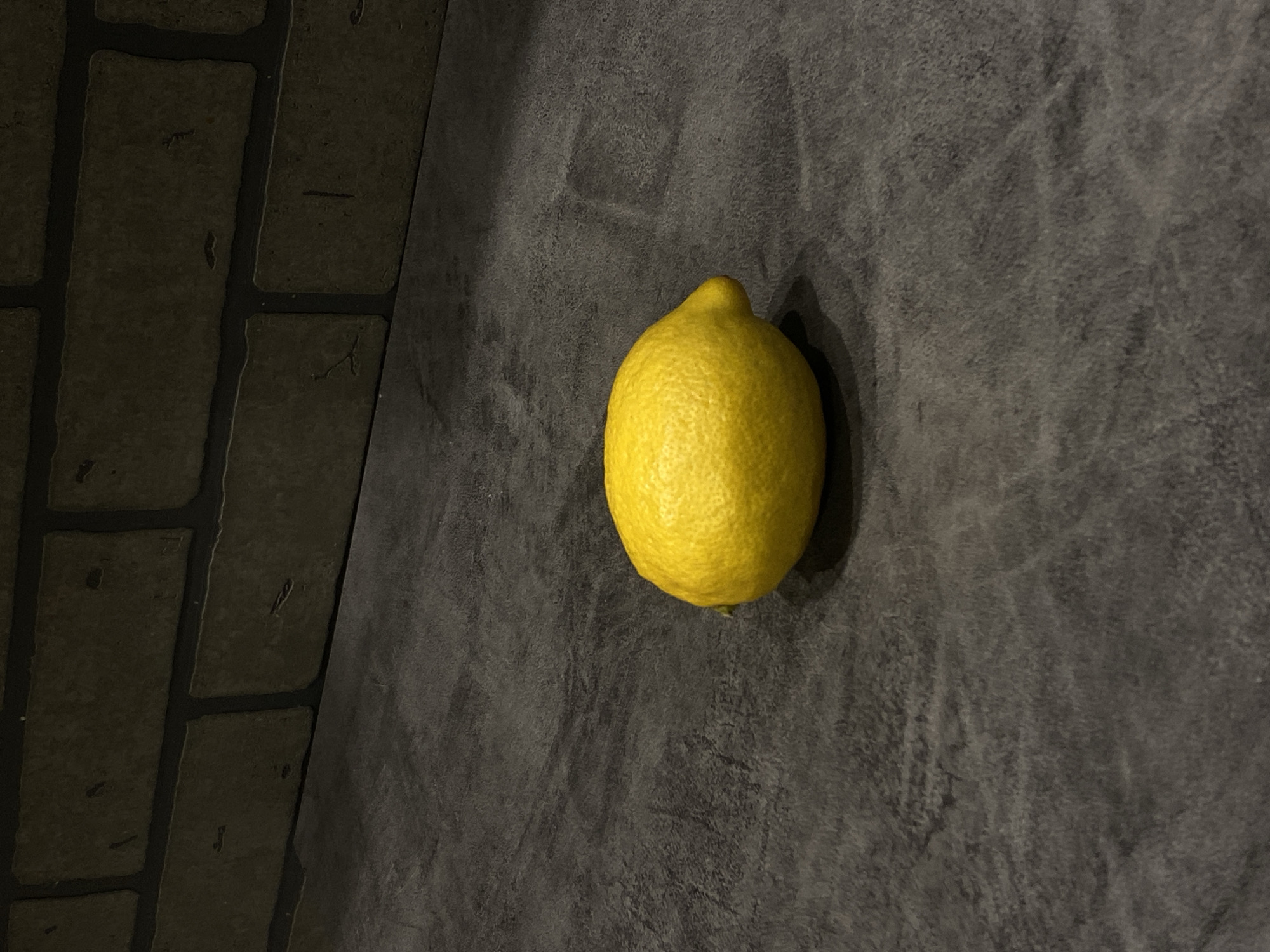 A lemon rests on a dark gray, textured surface, with a section of what appears to be brick visible in the lower left corner. The lighting is subdued, casting a shadow under the lemon. The environment appears indoors. There is no indication of Sochi, Russia.