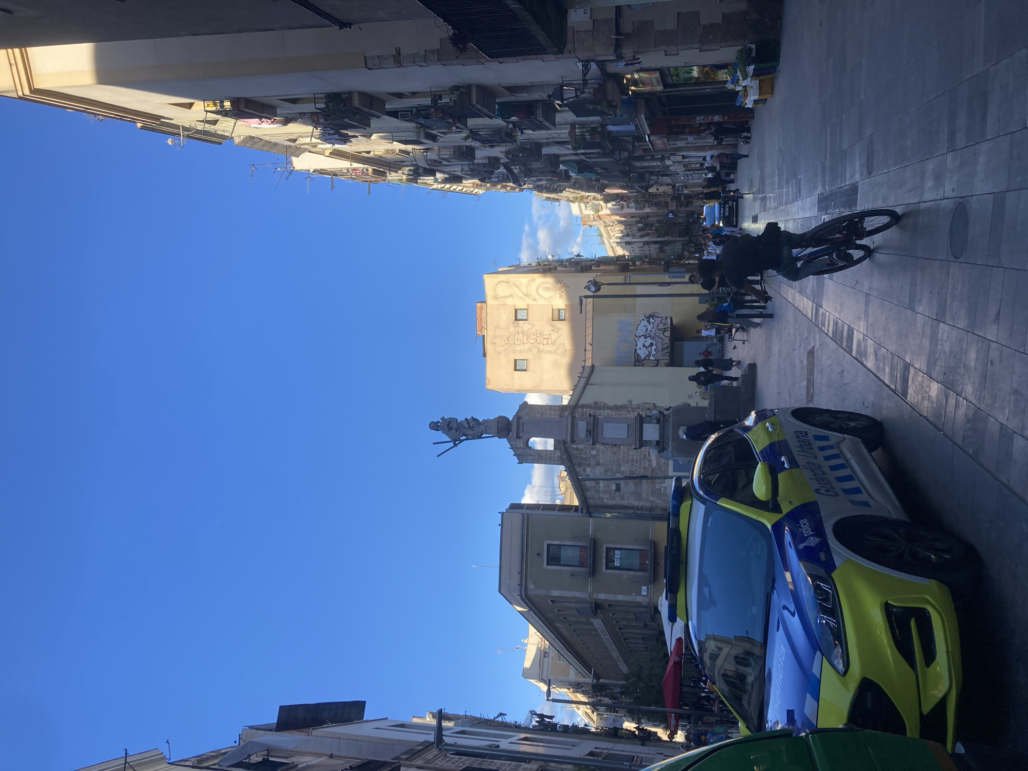 The image captures a street scene, likely in an urban setting within Unknown, Unknown. Dominating the foreground is a police car with a blue and yellow color scheme, partially obscuring another vehicle to its left. Further into the frame, a cyclist is seen riding on the pavement. Buildings line both sides of the street, rising several stories high and exhibiting varied architectural styles. In the middle distance, a monument or statue stands, suggesting a potentially historical or landmark location. The sky is clear and blue. People are visible in the middle ground, possibly engaged in casual activities.
