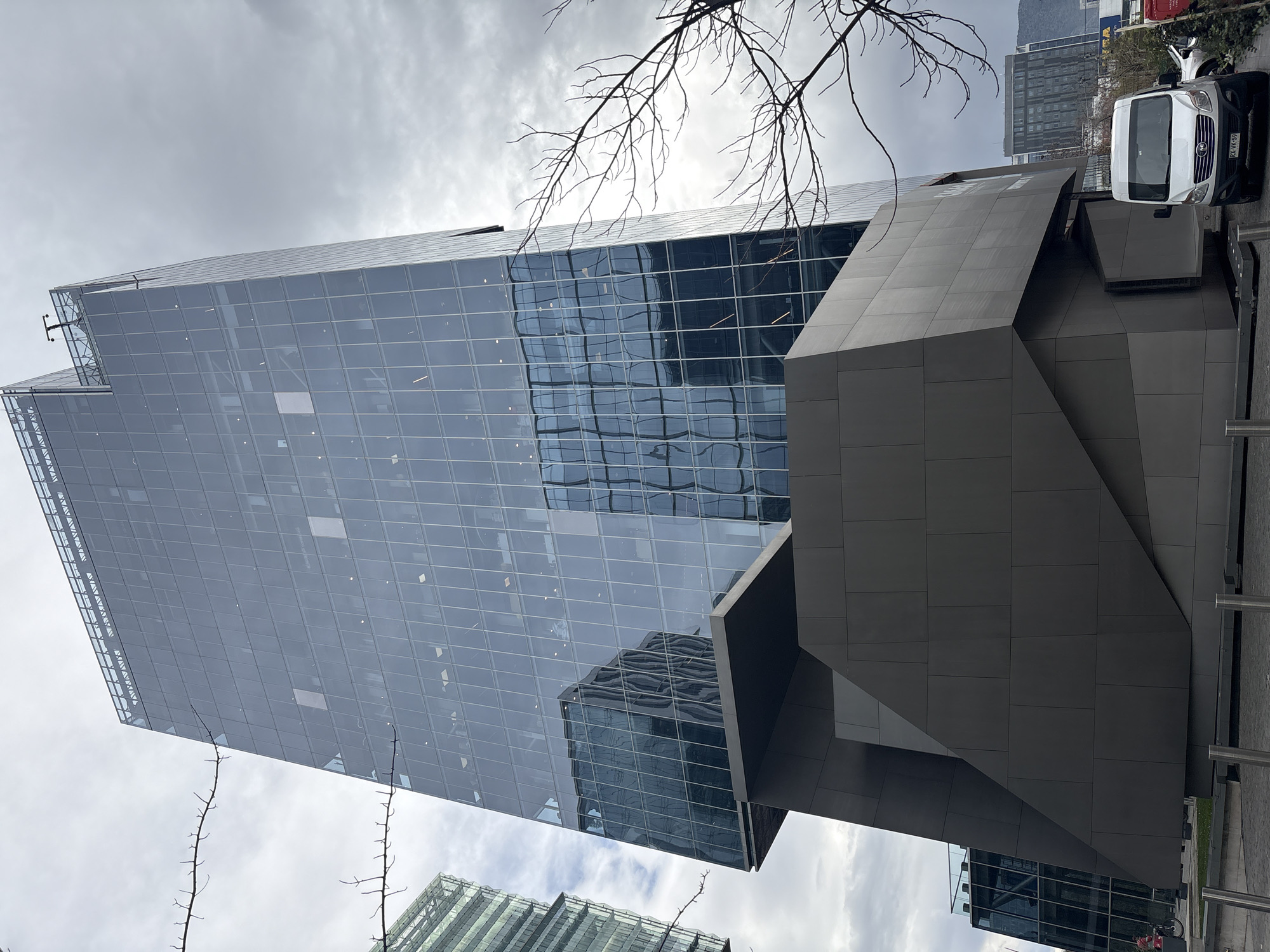 This is a ground-level, exterior, medium shot of a modern, glass-clad skyscraper in Santiago, Chile. The building features a geometric, angular design with a dark, faceted base. The sky is overcast. In the foreground, a white panel van with a license plate reading "RX VK-59" is parked on a paved area. Bare tree branches extend into the frame from the left and right. In the background, other buildings and distant hills are visible. A person is partially visible on the left, near a bicycle.