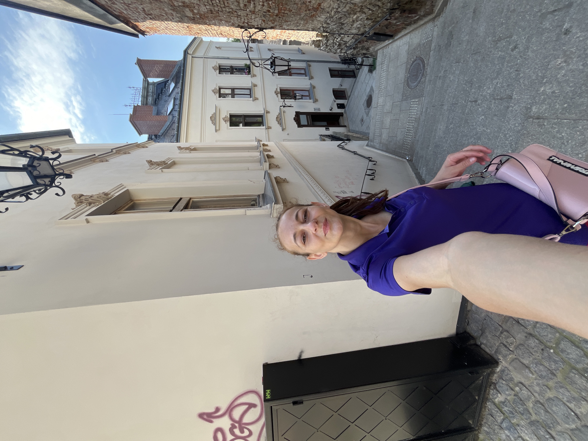 A woman, holding a phone out to take a selfie, stands on a cobblestone street lined with old buildings. She is wearing a purple polo shirt and a pink purse. The street is a steep staircase descending between the buildings. The buildings have ornate architectural details and some have graffiti on the walls. The sky is blue with some clouds. The scene is in an urban environment.