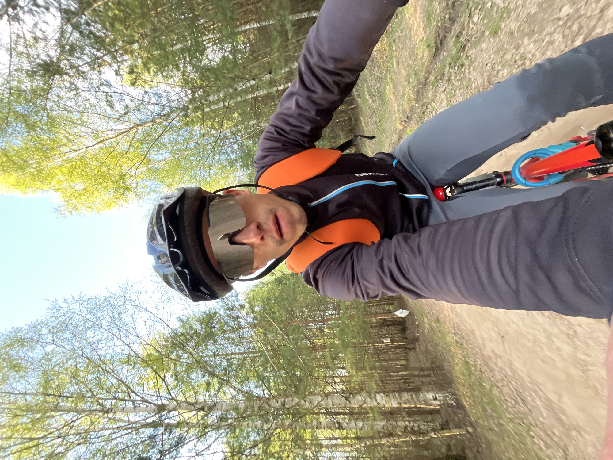 A man wearing a cycling helmet and sunglasses is riding a bike on a dirt path in a forest. He is wearing a black and blue long-sleeved shirt with orange straps. He is also wearing gray pants. The bike has red and blue accents. The background shows trees and foliage. The location is Chaikovsky, Russia, based on image context.