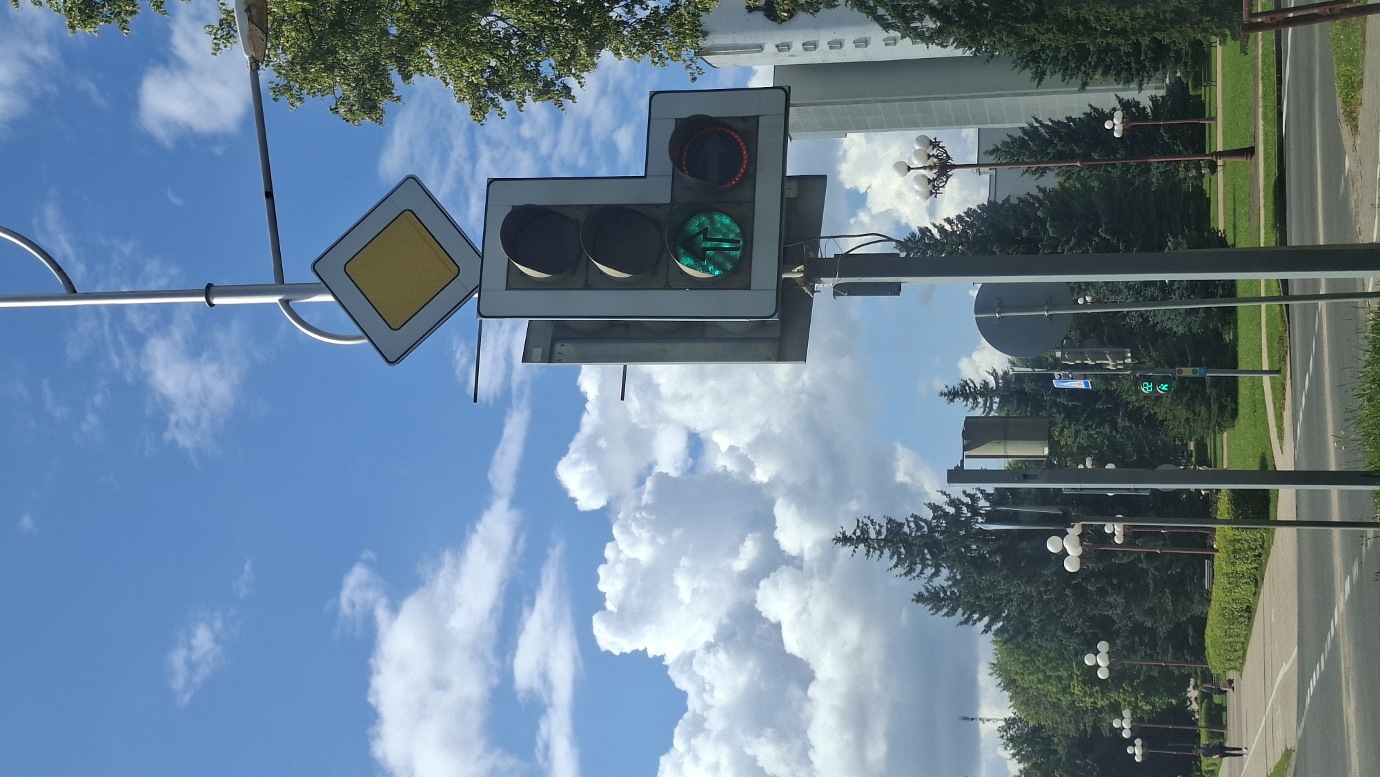 The image shows a traffic intersection in Maladzyechna, Belarus, under a blue sky with fluffy white clouds. A traffic light, displaying a green arrow pointing upwards and a red circular light, is mounted on a pole. Above the traffic light, a diamond-shaped road sign with a yellow center and a white border is visible. The surrounding environment includes manicured green hedges, tall evergreen trees, lampposts with multiple globes, and a modern building. A paved walkway with a bike lane runs alongside the road. A person is walking on the walkway in the distance, and another person appears to be sitting on a bench. The road surface in the foreground shows white lane markings.