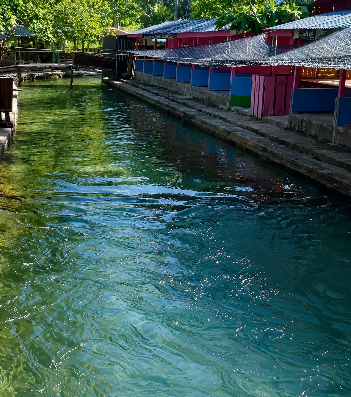 This image captures a serene canal in Silaiyajae, Indonesia. The clear, turquoise water flows gently, reflecting the lush green foliage and the bright blue sky. Along the right bank of the canal are several open-air structures with pink roofs and blue and green wooden partitions, suggesting they are possibly food stalls or rest areas. A wooden walkway with a railing is visible on the left side of the canal, leading to what appears to be a small bridge. The scene is tranquil and appears to be a popular spot for relaxation and enjoying nature.