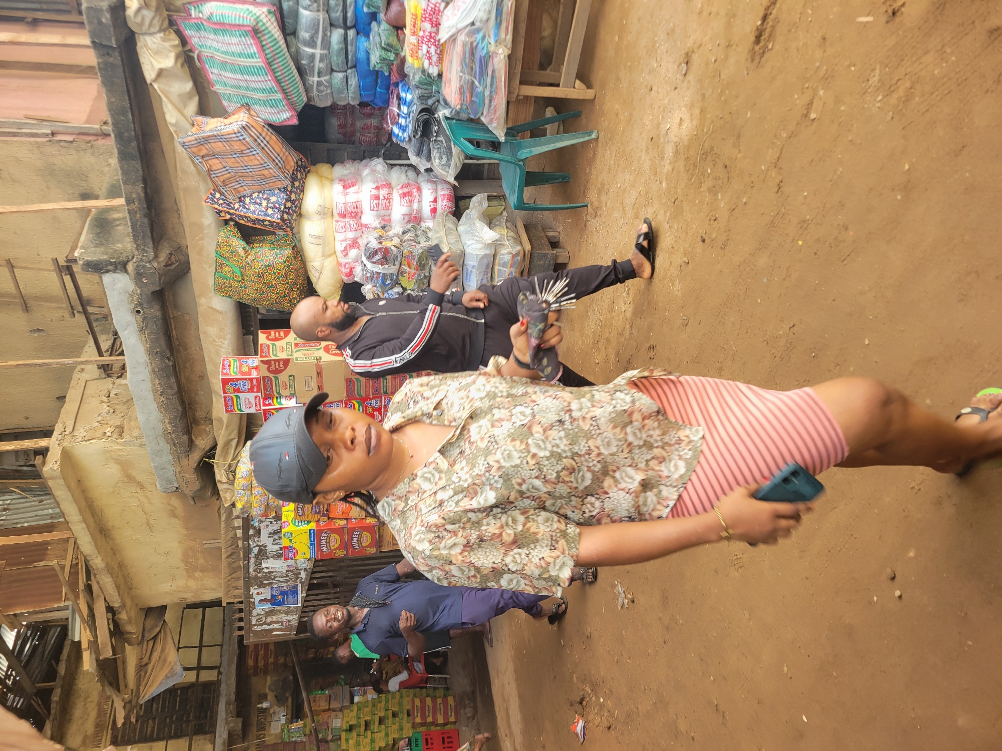 A medium shot depicts a woman in a floral blouse, pink striped skirt, and baseball cap, holding a smartphone and facing the camera. Behind her, a man in a tracksuit reclines amongst stacked merchandise, while another man in a blue shirt is visible. The background features a cluttered roadside market in Onitsha, Nigeria, including various goods such as packaged items, bags, and plastic chairs. The ground is dirt.