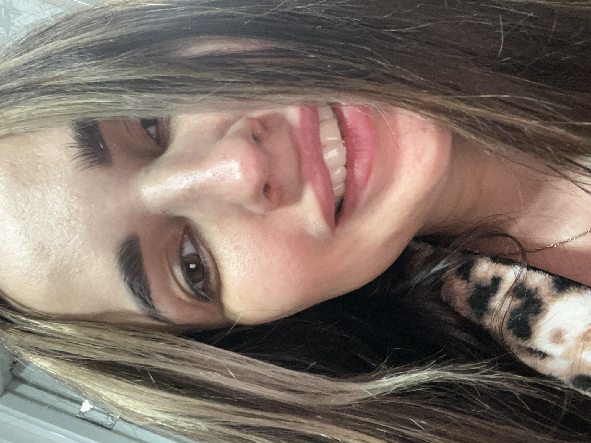 A close-up, eye-level selfie depicts an adult female smiling directly at the camera. The subject has long, brown hair with light blonde highlights, falling over her shoulders and partially obscuring the sides of her face. Her eyes are brown, and her eyebrows are dark and full. She wears light pink lip color and displays her upper teeth in a smile. The skin on her face exhibits a light flush on the cheeks and nose. A portion of a brown, black, and tan leopard print garment is visible on the subject's left side (viewer's right), and a thin, gold-colored chain necklace is visible on her right side (viewer's left). The background is indistinct, showing minimal details of an indoor setting, including a light-colored surface and a faint, indeterminate object in the upper left. The specific location in the city of Unknown, Unknown, cannot be determined from the image.