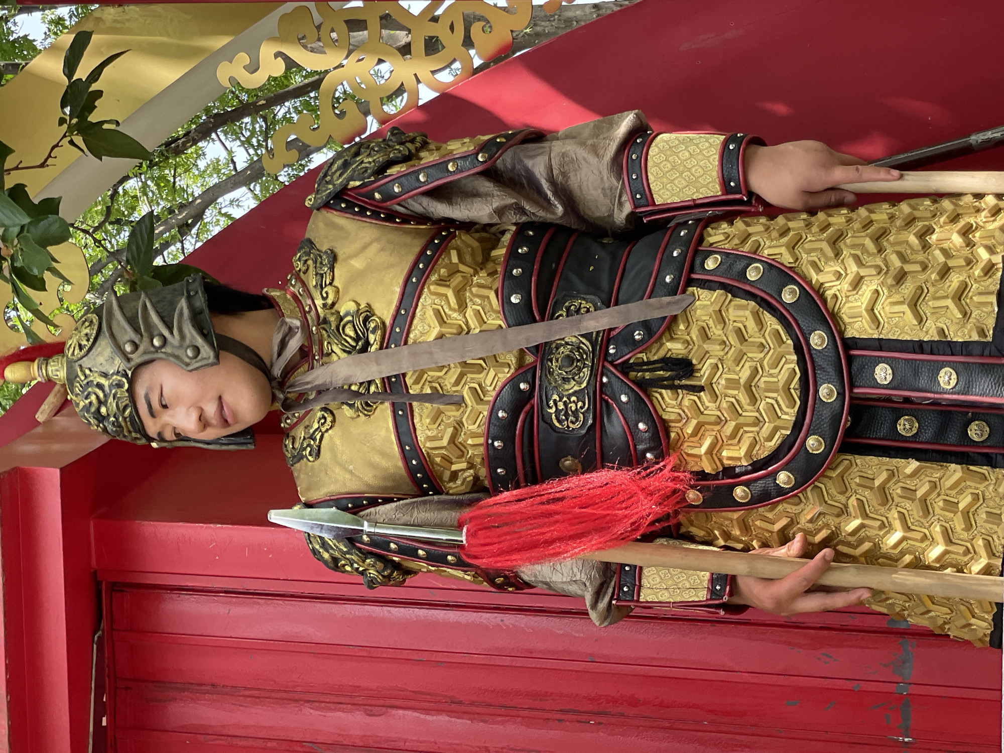 A person in golden Chinese armor stands guard in Lintong District, China. The individual wears a helmet and holds a spear. The backdrop is a red wall and an ornate golden decoration.