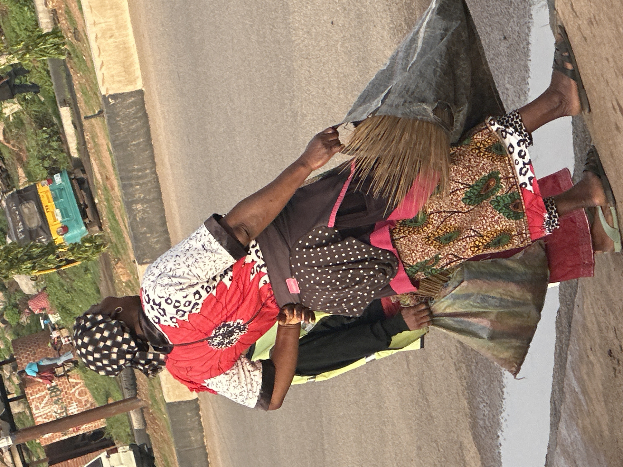 In Gura Top, Nigeria, a person is walking on the side of a road. They are wearing a red and white patterned shirt and a dark skirt with white polka dots. A large, woven basket is held in their right hand. To their left, another person is visible, partially obscured, wearing a yellow vest over a black long-sleeved shirt. In the background, a small, green and yellow three-wheeled vehicle is parked on a grassy embankment. The road is paved, with a concrete curb and a grassy area beyond. The overall scene suggests an outdoor, everyday environment in a Nigerian city.