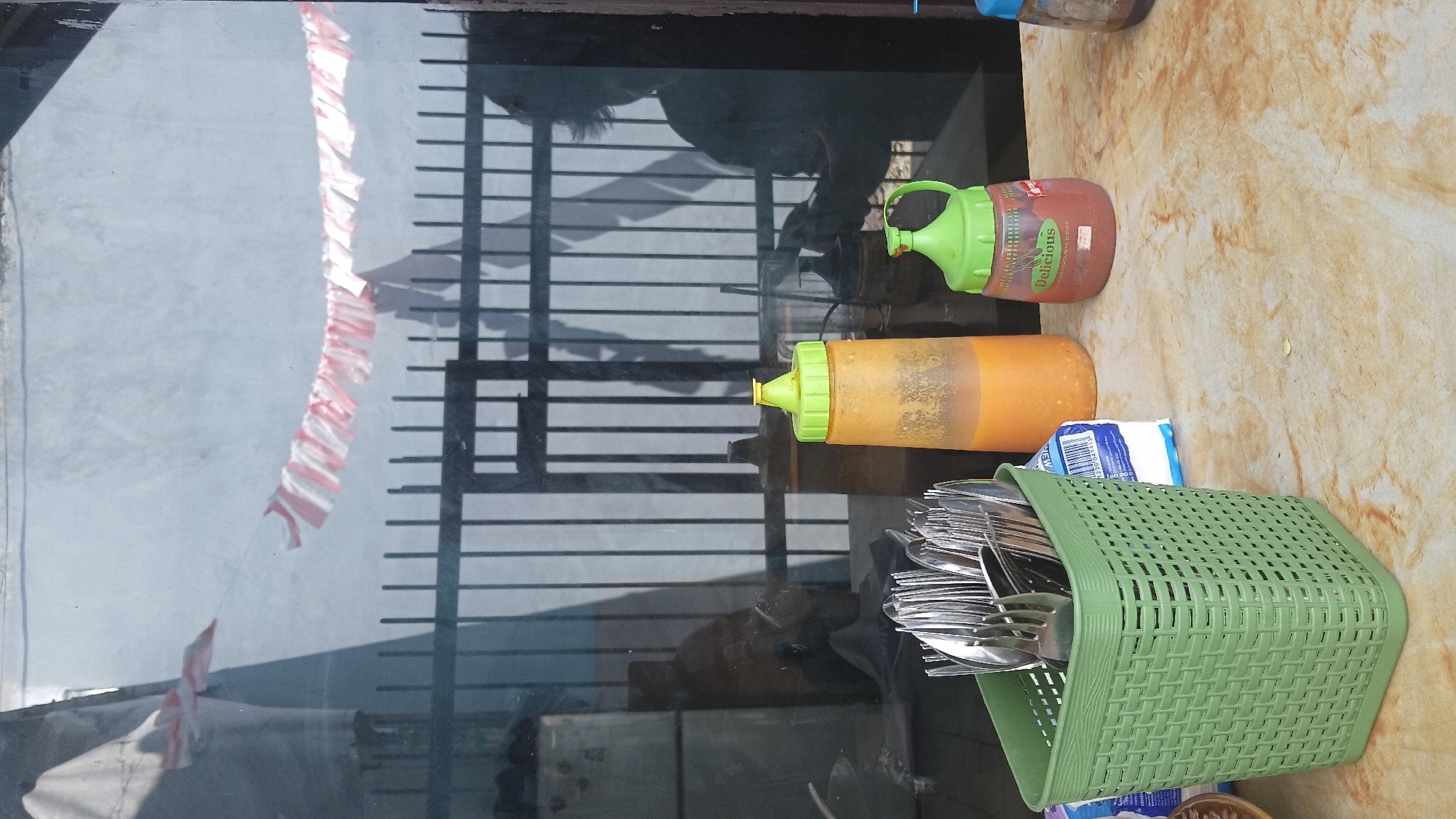 The image depicts a tabletop scene with various items, likely at an eatery in Dayeuhkolot, Indonesia. A green woven basket on the left holds a bundle of silverware, primarily spoons and forks. To the right of the basket are two squeeze bottles, one orange with a yellow cap and the other red with a green cap. The red bottle has the label "Delicious" on it. Behind the bottles, a reflection in a window shows vertical bars of a fence and a person's silhouette. In the background, a string of red and white flags, possibly for a national celebration, is draped across the window. The tabletop itself has a marbled texture. A white package, possibly of napkins or tissues, is visible between the basket and the orange bottle.