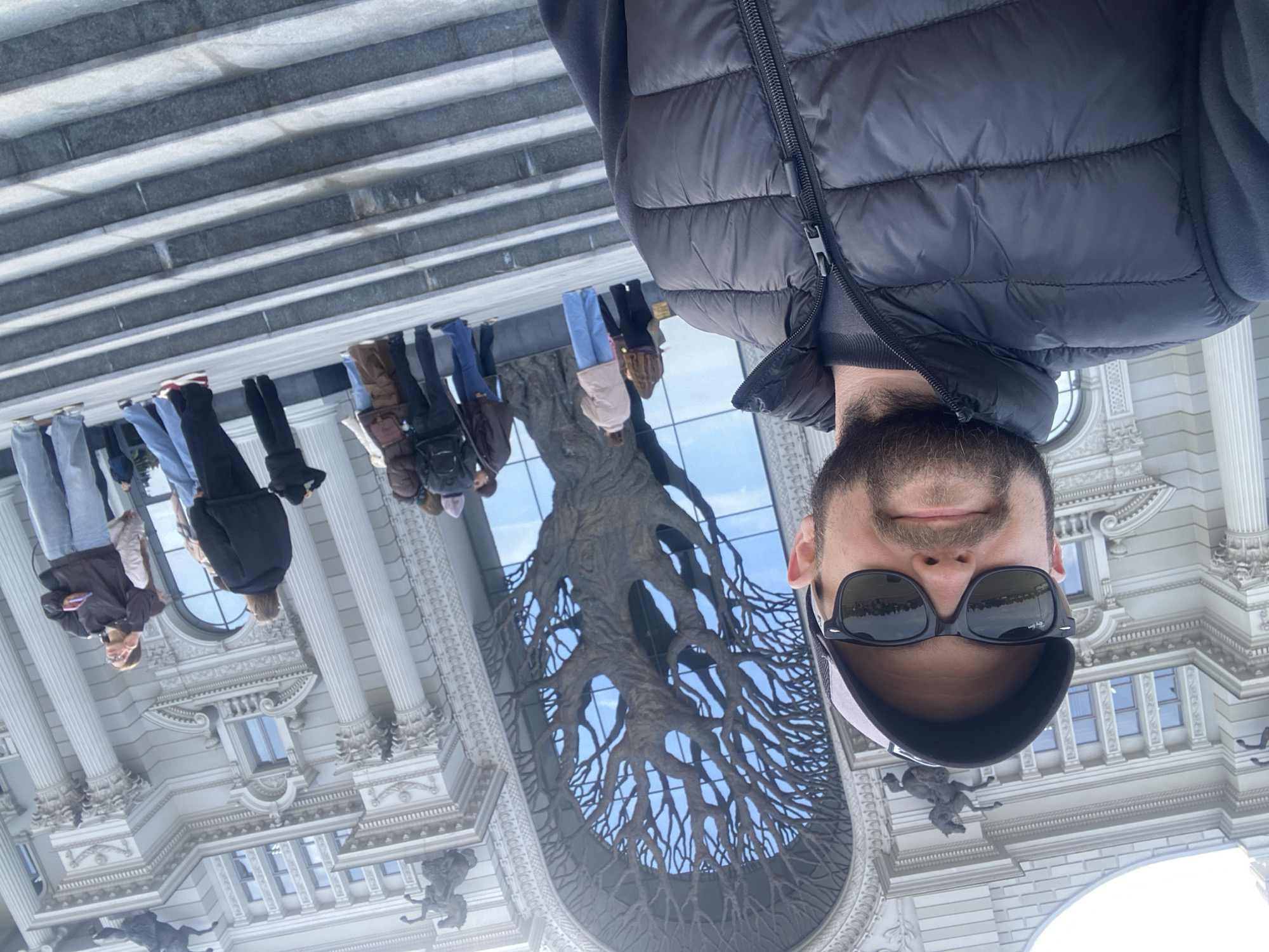 A man with a beard and sunglasses, wearing a black puffer jacket and cap, is the primary subject in this selfie-style photograph. He is centered in the frame, and the perspective is slightly angled upward.
Behind the man, there is a large, ornate building. The building features a tall arched entrance with a decorative sculpture of a tree made of metal within the archway. The building is adorned with architectural details such as columns, windows, and decorative sculptures. Several people are walking or standing near the building.
The location is the Palace of Farmers in Kazan, Russia. The large metal tree sculpture is a prominent feature of the building's facade.