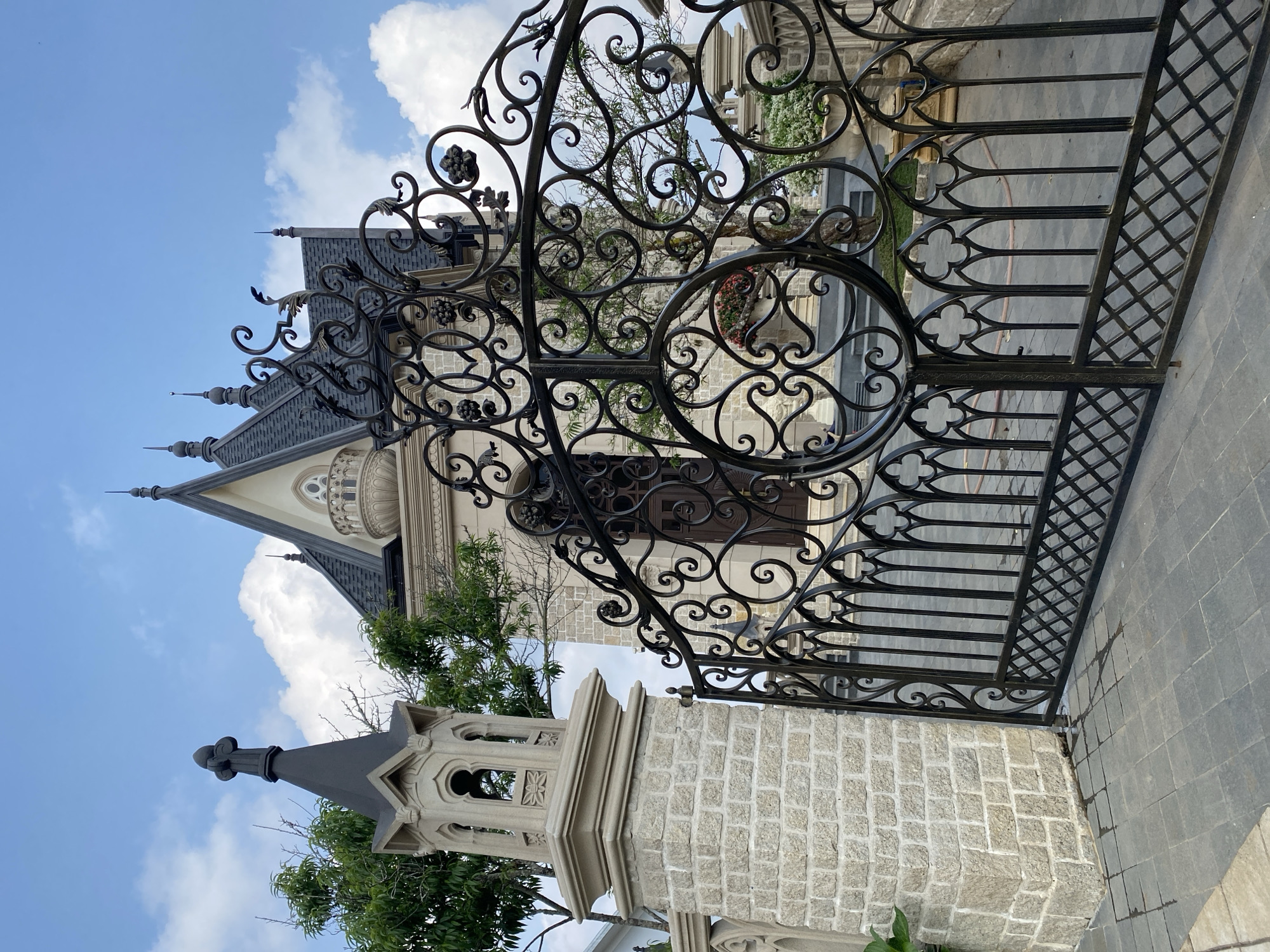 The image presents a low-angle view of a wrought iron gate, partially open, revealing architectural elements of a European-style building in the background. The gate's intricate design features decorative swirls, floral motifs, and vertical bars with trefoil detailing. The building behind the gate exhibits a steeply pitched, slate-gray roof with pointed spires, suggesting a castle or church-like structure. The facade is a combination of light-colored stone and stucco. A stone turret or tower is visible to the left of the gate, constructed from light gray blocks and topped with a similar spire. The sky is blue with scattered white clouds. The location is recognizable as being in Da Lat, Vietnam, known for its European-inspired architecture.