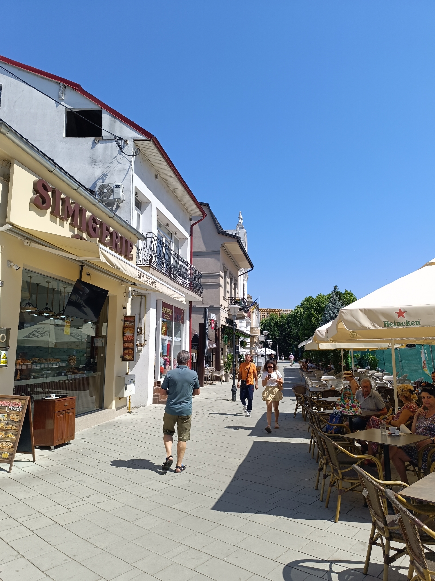 This image captures a street scene in Târgu Jiu, Romania, likely on a sunny day. The perspective is from the ground level, looking down a pedestrianized street lined with shops and outdoor cafes.
On the left, a building facade features a shop with large glass windows displaying baked goods. Above the windows, the sign "SIMIGERIE" is prominently displayed. An air conditioning unit is visible on the building's exterior. A wooden display stand with a menu featuring pictures of food items is placed on the sidewalk in front of the shop.
The sidewalk is paved with square gray tiles. Several people are walking along the street. In the foreground, a man in a blue t-shirt and khaki shorts is walking away from the camera. Further down the street, a couple is walking towards the camera; the woman is wearing a white top and a yellow skirt, and the man is wearing an orange t-shirt and dark pants.
On the right side of the street, a row of outdoor tables and chairs are set up under large beige umbrellas, some bearing the "Heineken" logo. Several patrons are seated at these tables, engaged in conversation and dining. The background shows more buildings, trees, and a clear blue sky. The overall atmosphere suggests a lively commercial area with opportunities for dining and shopping.