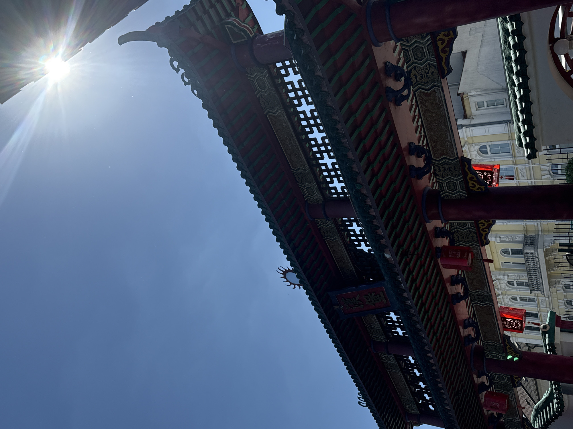 This upward-angled photograph captures a large, ornate Chinese-style gate (Paifang) against a bright blue sky. The gate features traditional architecture with prominent red pillars, intricate green and red tiled roof structures, and decorative elements. Numerous red lanterns with delicate patterns hang from the gate's beams.
The sun is visible in the upper left corner, casting a strong flare and radiating beams of light across the clear sky, indicating bright daylight, likely midday or early afternoon. Tiny dust particles are illuminated by the strong sunlight in the sky.
Through the intricate lattice work and gaps in the gate's structure, parts of a light-colored, multi-story European-style building are visible in the background, featuring arched windows and decorative facades.
Visible text on a red plaque on the gate includes two Chinese characters, which appear to be "廣榮" (Guǎng Róng), often meaning "extensive glory" or similar auspicious connotations.
The scene depicts a unique cultural landmark under a clear, sunny sky, creating a contrast between the ornate Chinese gate and the underlying European urban architecture. Given the location hint, this image is from Hainburg an der Donau, Austria.